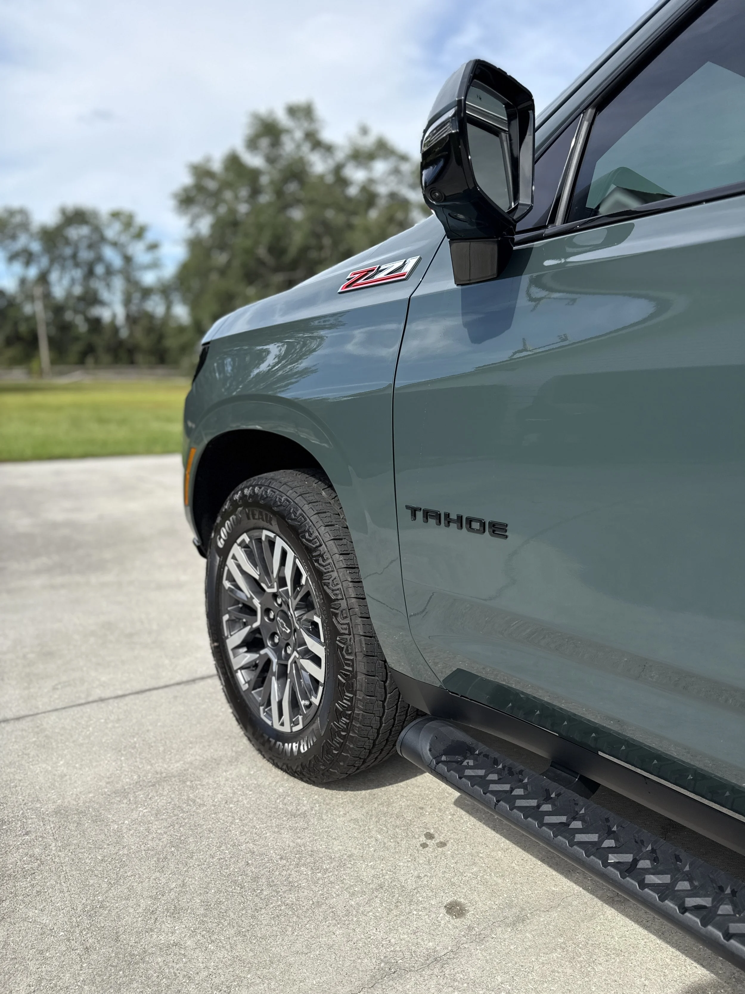 Close-up of a gray Chevrolet Tahoe SUV showcasing the front passenger side, side mirror, tire, and Z71 emblem with a green landscape and blue sky in the background.