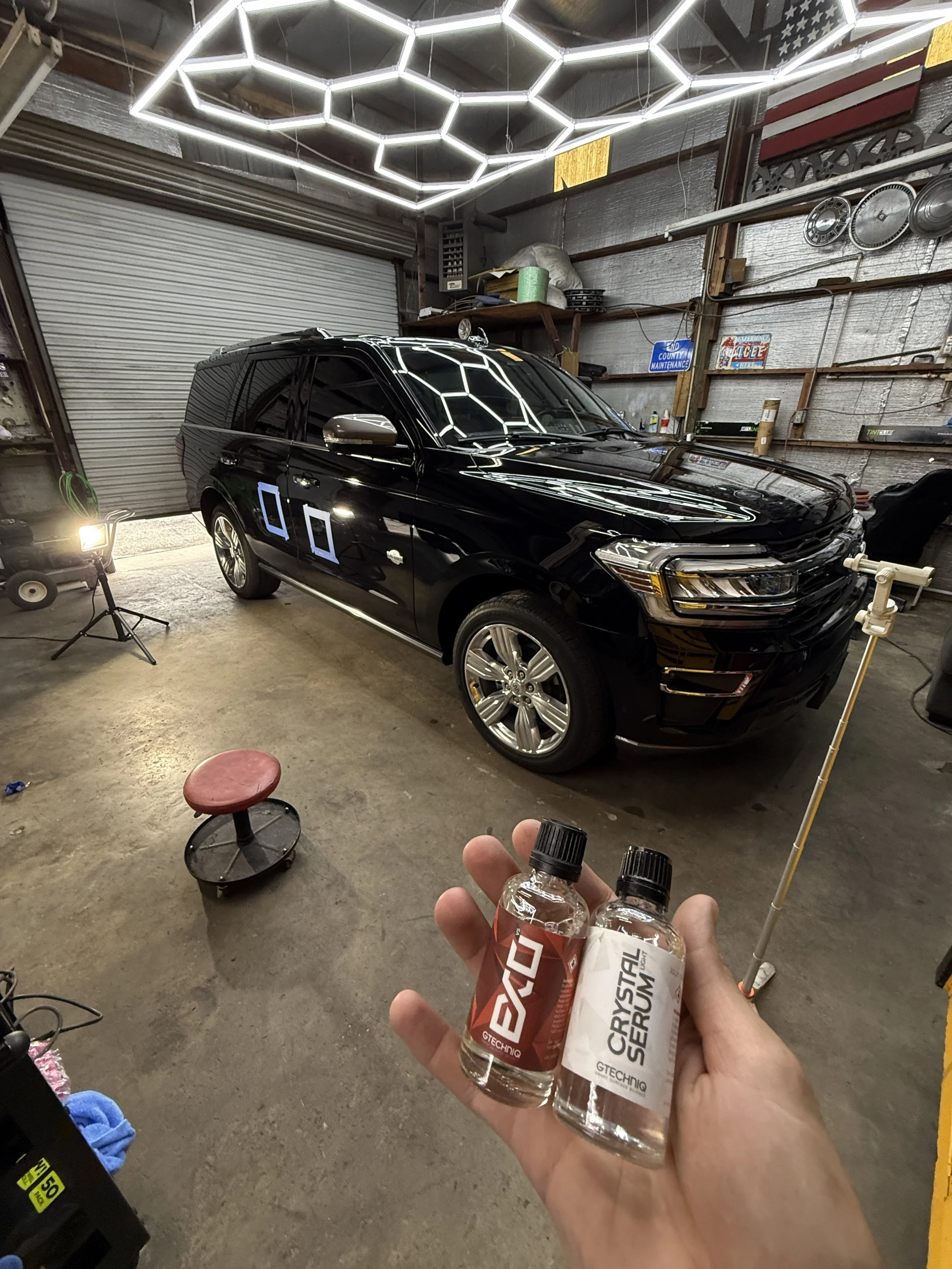 A person holding two small bottles labeled 'CRYSTAL SERUM' and 'GTECHNIQ' inside a garage with a black SUV parked inside. The garage has metal walls, shelves with various items, workout equipment, a stool, and ceiling lights arranged in a honeycomb p