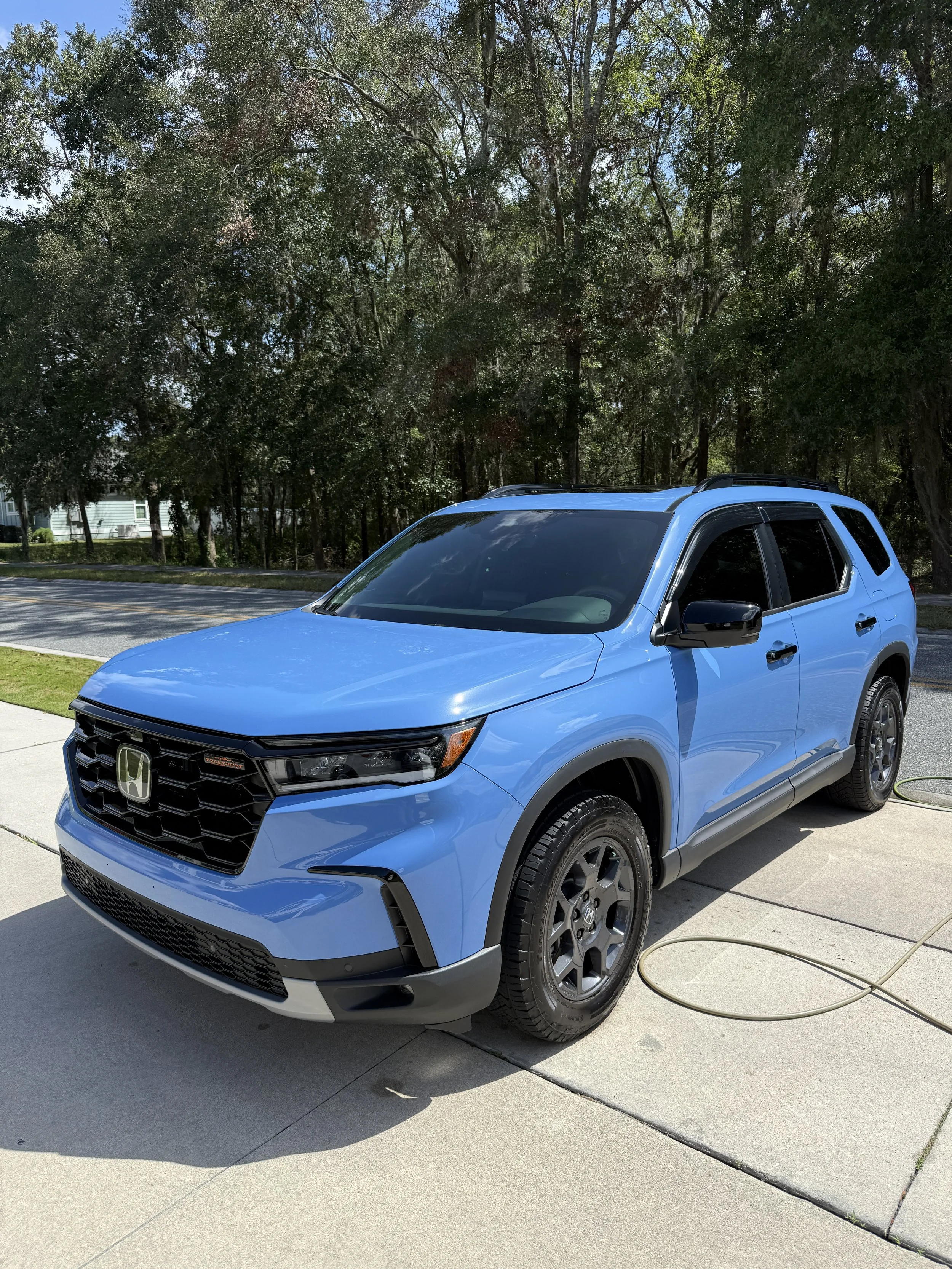 Blue Honda SUV parked on a driveway next to a garden hose with a background of trees and a street.