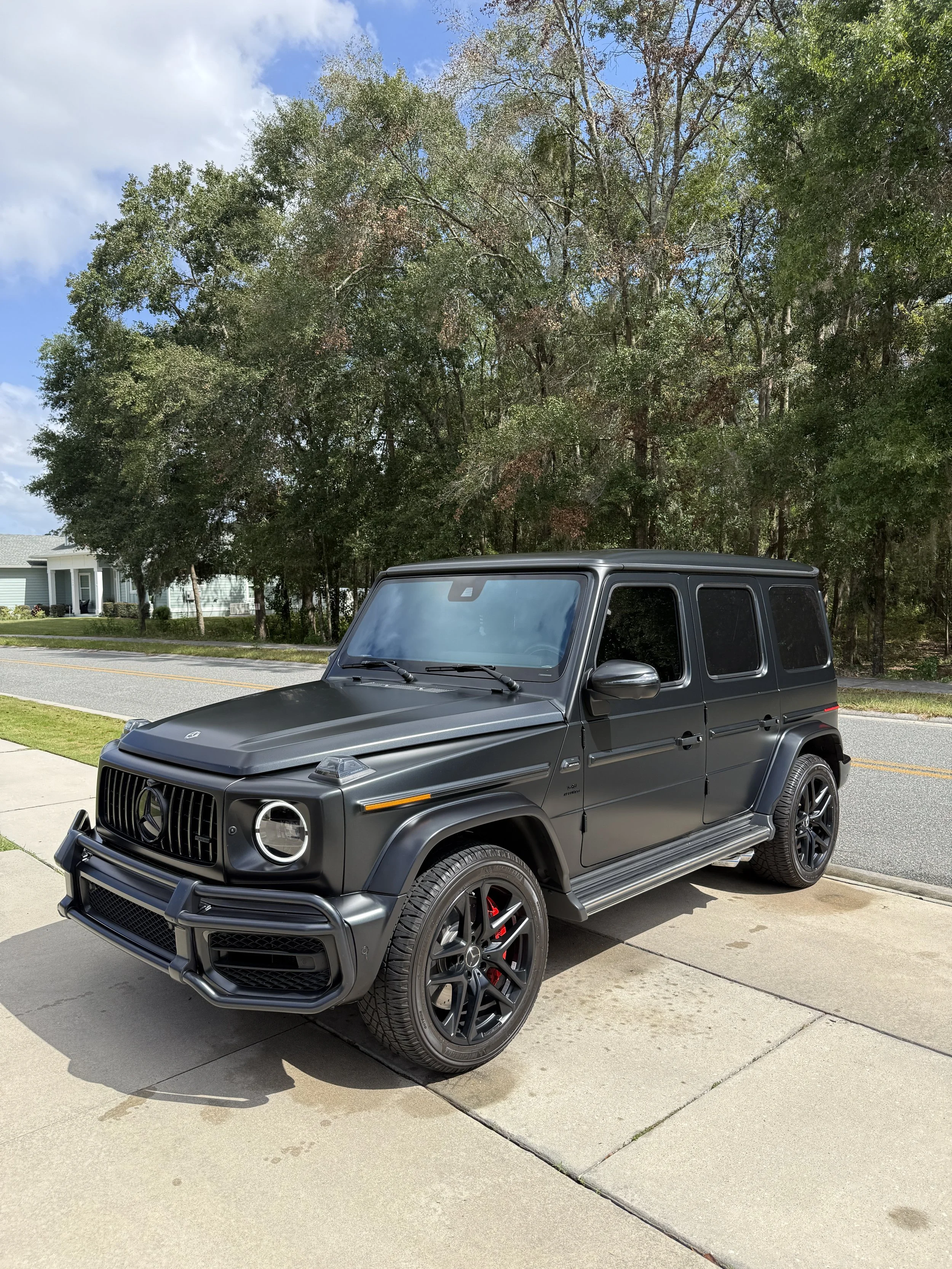 A black Mercedes-Benz G-Class SUV parked on a concrete driveway in front of a green yard and trees, with a blue sky and some clouds overhead.