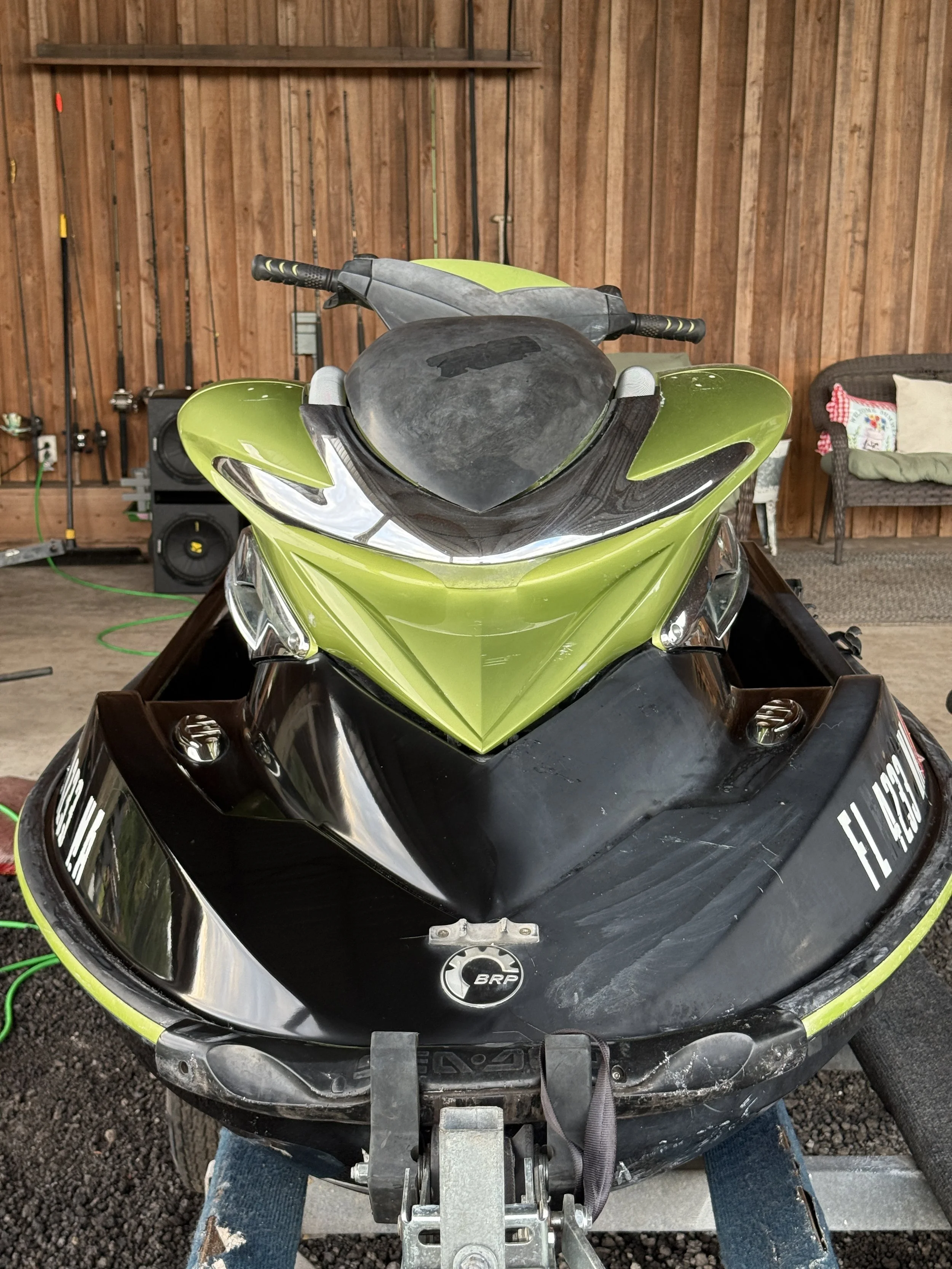 Green and black jet ski on a trailer in a wooden garage, with fishing rods on the wall and a speaker in the background.