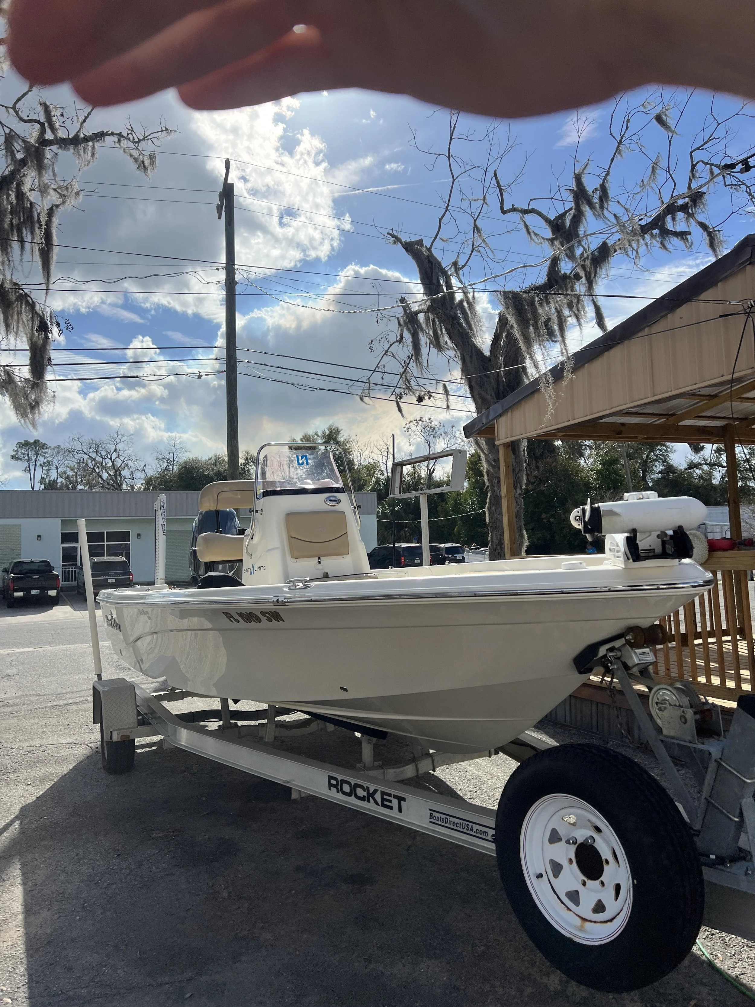 A small white boat on a trailer parked outside on a gravel lot, with a beige cushioned seat at the helm and electrical equipment attached, under a partly cloudy sky, with leafless trees and a building in the background.