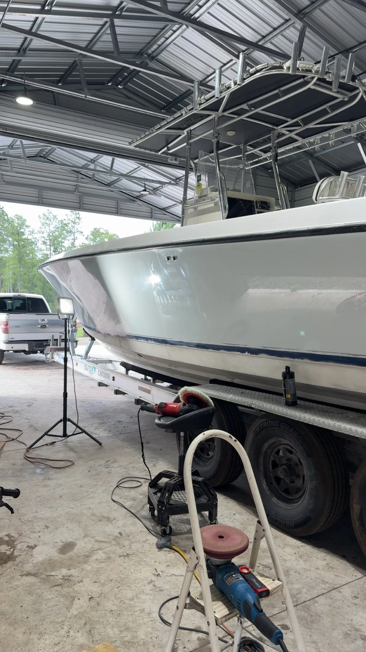 A white boat on a trailer inside a large garage or workshop, with tools and equipment around, including a work light, angle grinder, and a small step ladder.