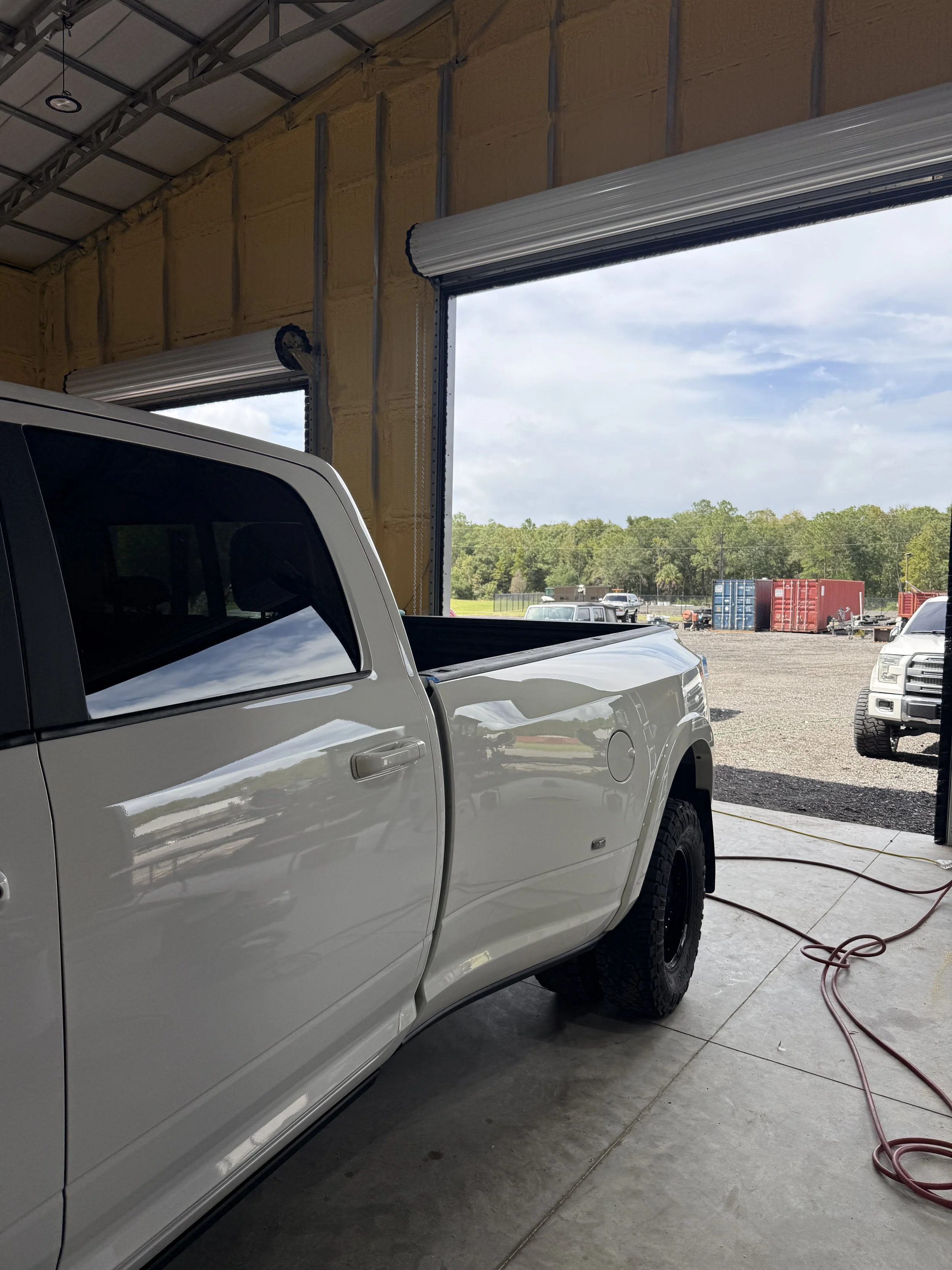 Inside a garage with a white pickup truck, partially visible, parked near the open garage door. Outside, there are other vehicles, storage containers, and trees in the background.