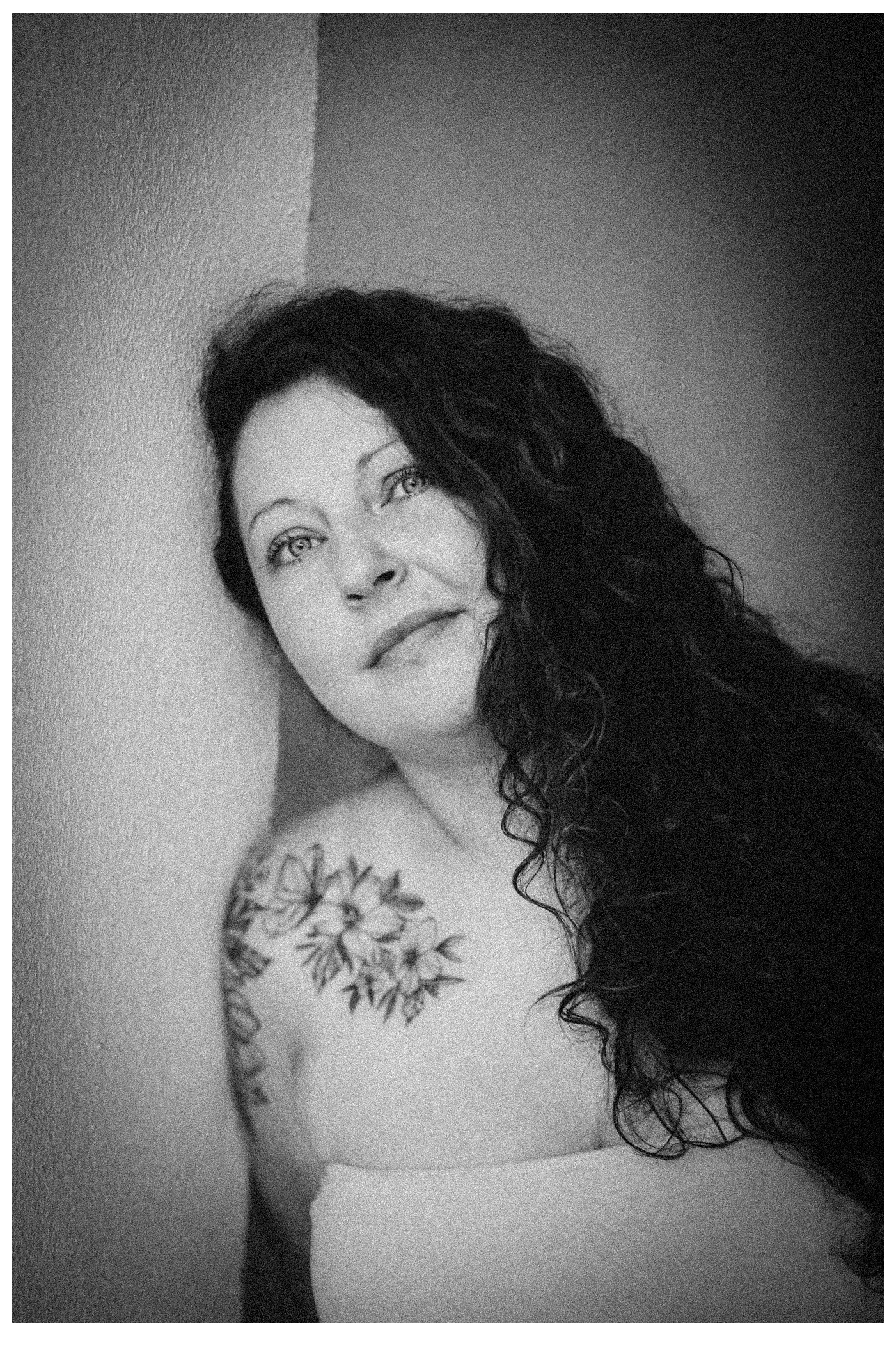 Schwarz-weiß Porträt einer Frau mit langen, lockigen Haaren, die an die Wand gelehnt ist, mit einer Blumentattoo auf der Schulter, blickt nach oben.