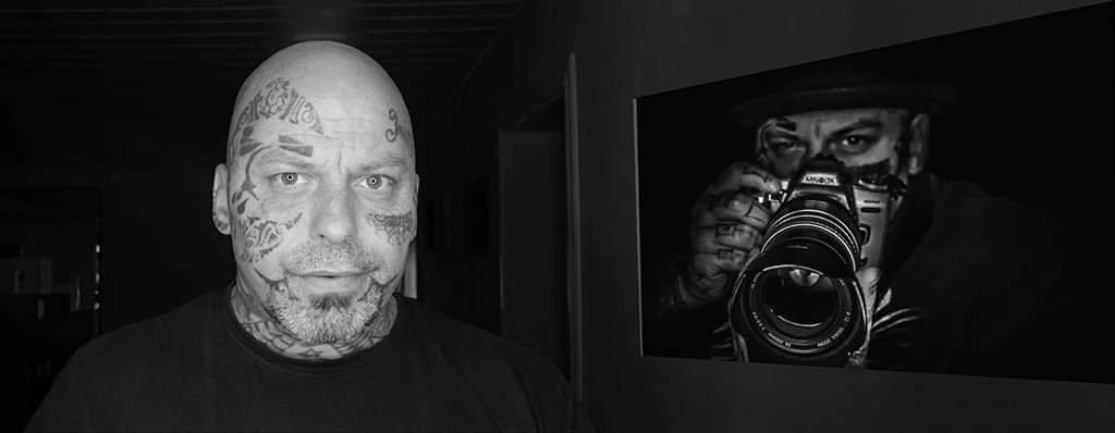 Schwarzweiß-Porträt eines Fotografen mit tätowiertem Gesicht vor einem Foto mit Kamera