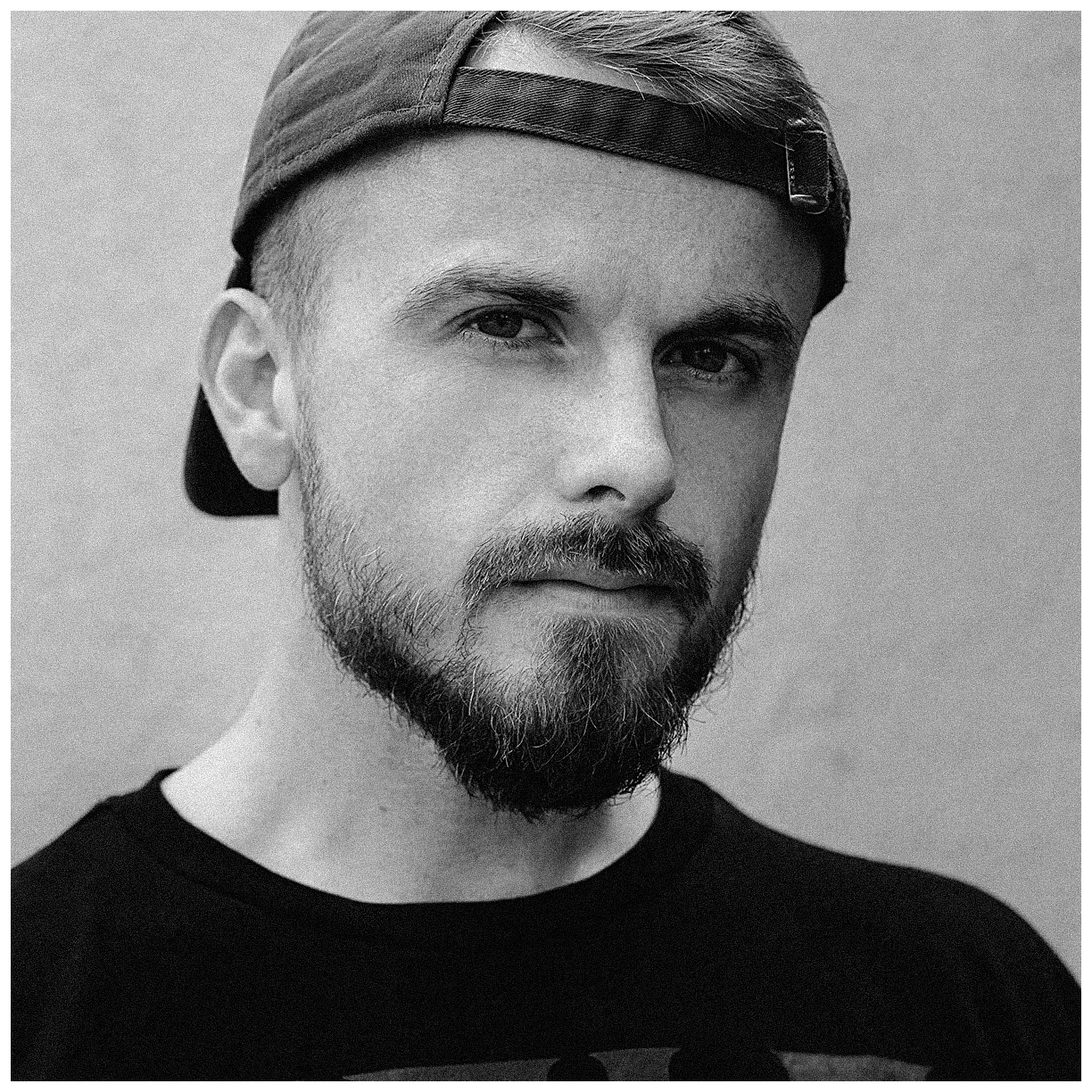 Schwarz-Weiß-Porträt eines jungen Mannes mit Bart, trägt eine schwarze Cap rückwärts und ein schwarzes T-Shirt, blickt ernst in die Kamera.
