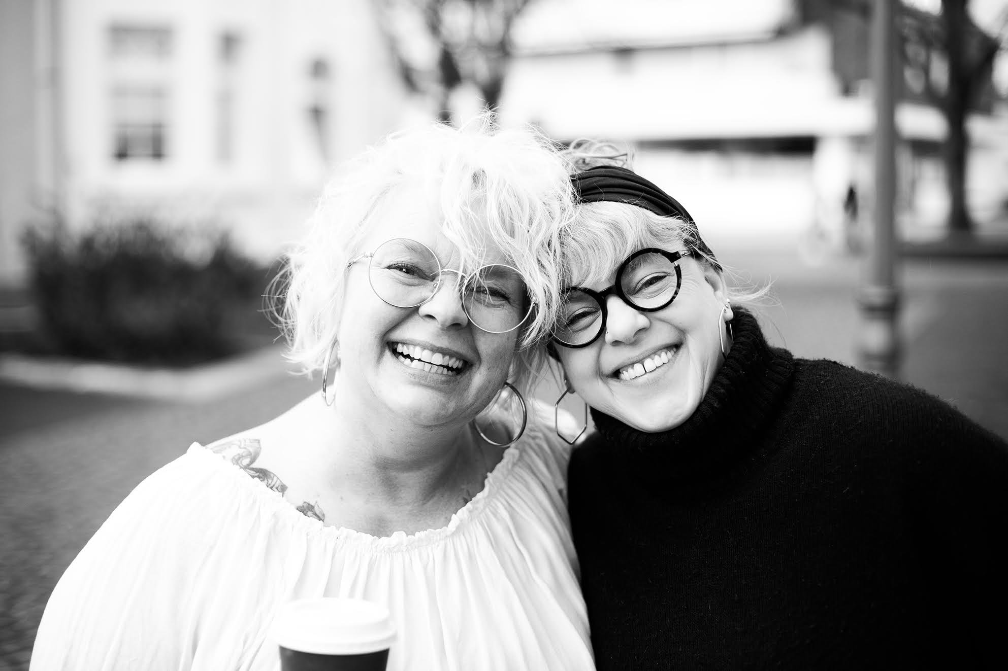 Zwei lachende Frauen im Freien, eine mit roten Haaren und große Brille, die andere mit kurzen Haaren, schwarze Kleidung und runde Brille, posieren für ein Selfie.