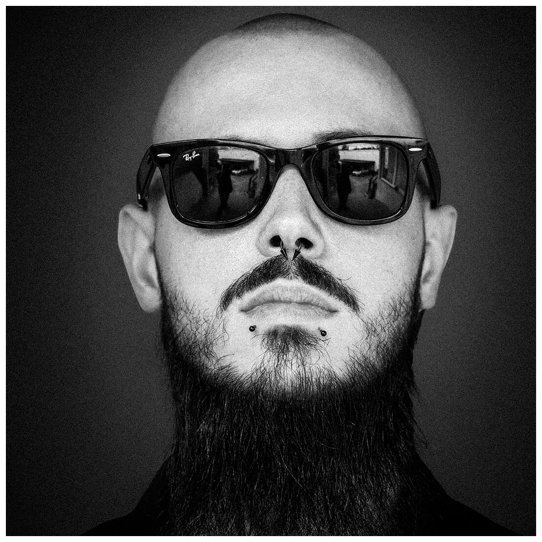 Schwarzer und weißer Porträtaufnahme eines Mannes mit Glatze, Bart und Piercings, der dunkle Sonnenbrille trägt. Die Brille reflektiert einen Raum mit Fenstern und Menschen.