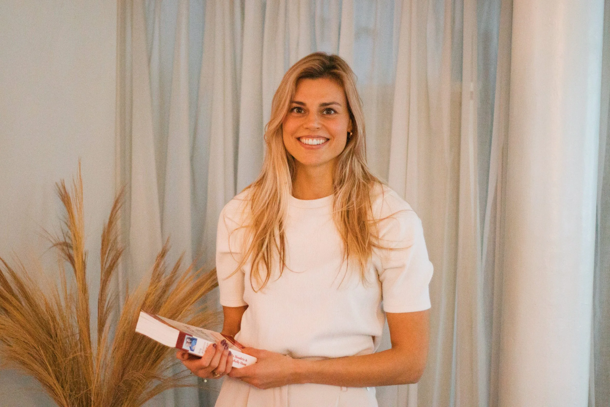 Een glimlachende vrouw met lang, golvend blond haar en een witte top, die binnen staat voor een achtergrond van lichtgekleurde gordijnen. Ze houdt een brochure of boekje vast in haar handen.
