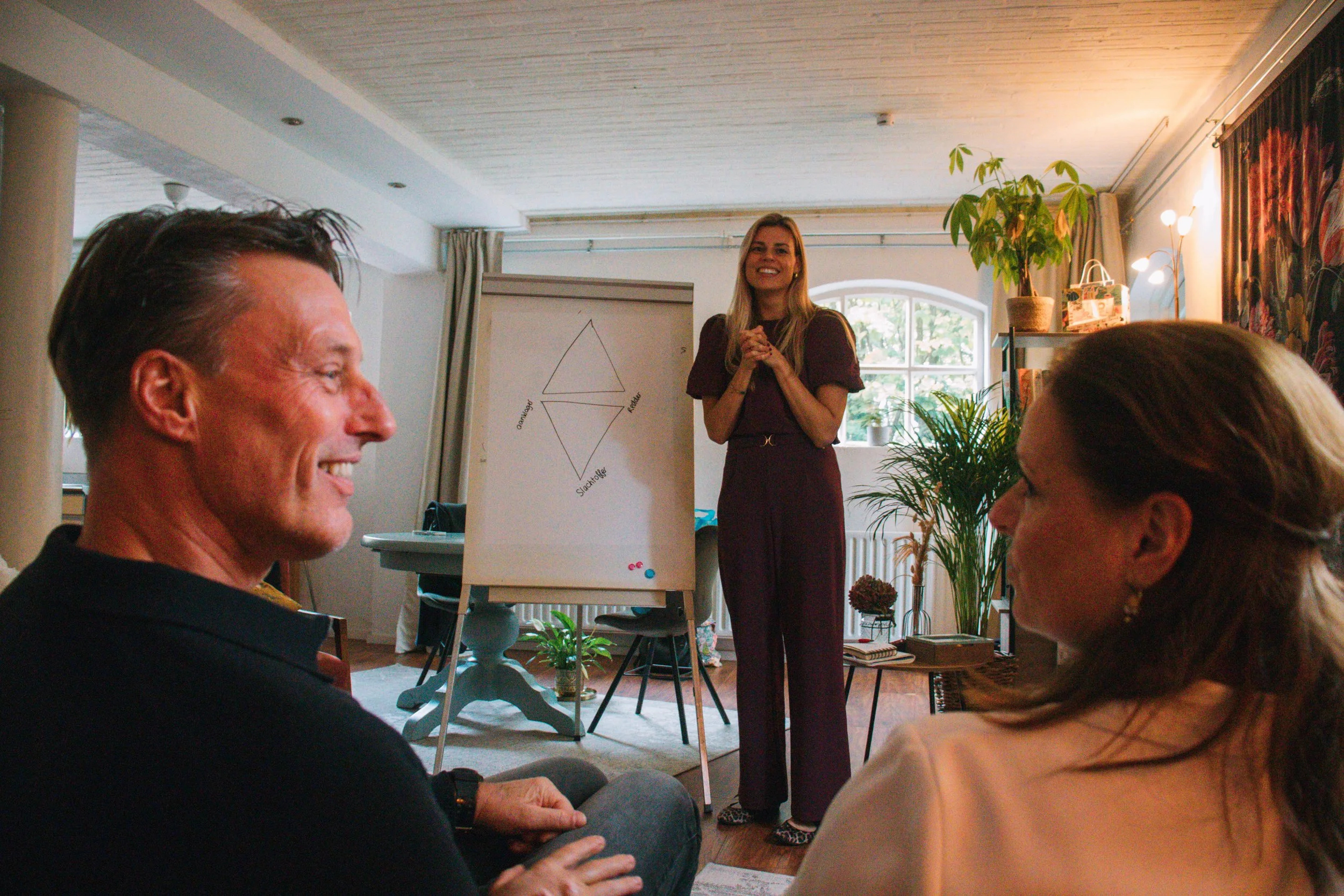Vrouw geeft presentatie in een kamer met grote ramen, groene planten en een witte muur met een poster met een diagram.