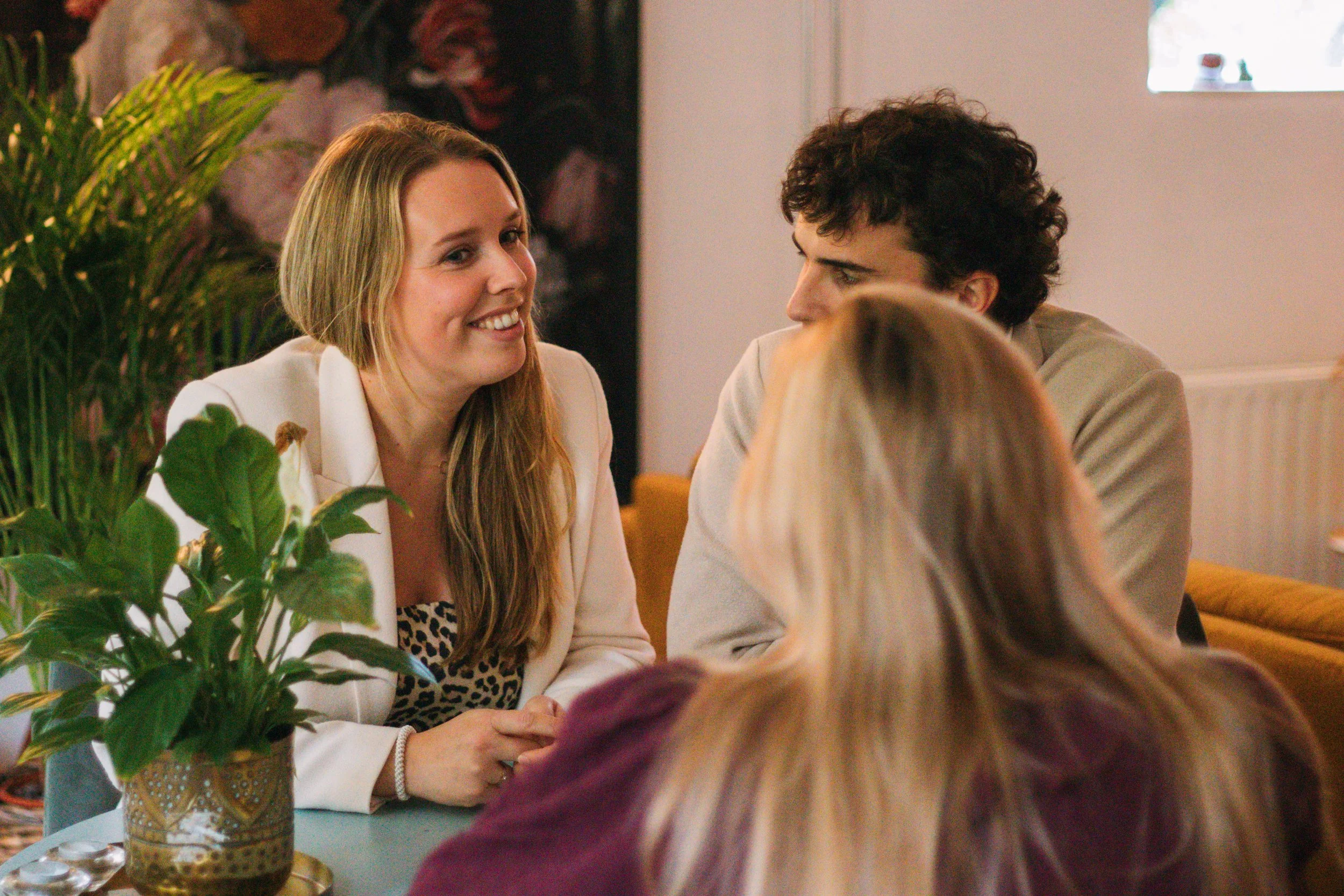 Een vrouw met lang haar die glimlacht en een man met krullend haar die naar haar kijkt, terwijl ze samen aan een tafel zitten in een gezellige setting. Op de voorgrond een plant in een gouden pot.