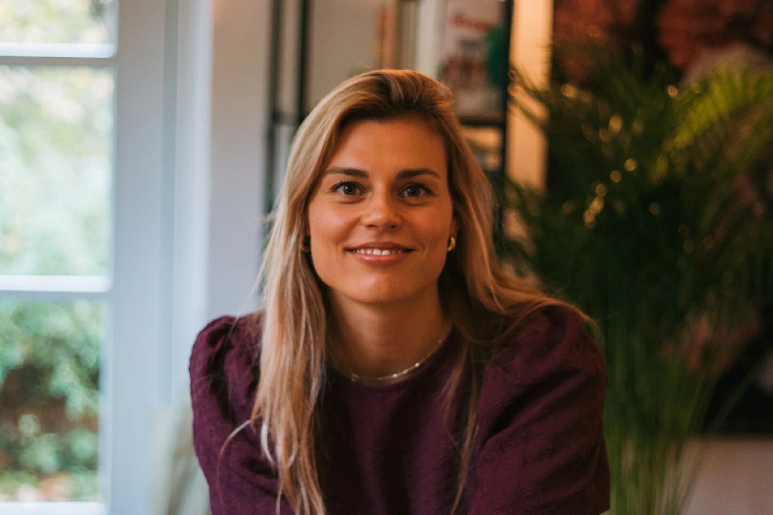 Een glimlachende vrouw met lang blond haar en een paar oorbellen, zittend binnen in een kamer met veel groen en een raam op de achtergrond.