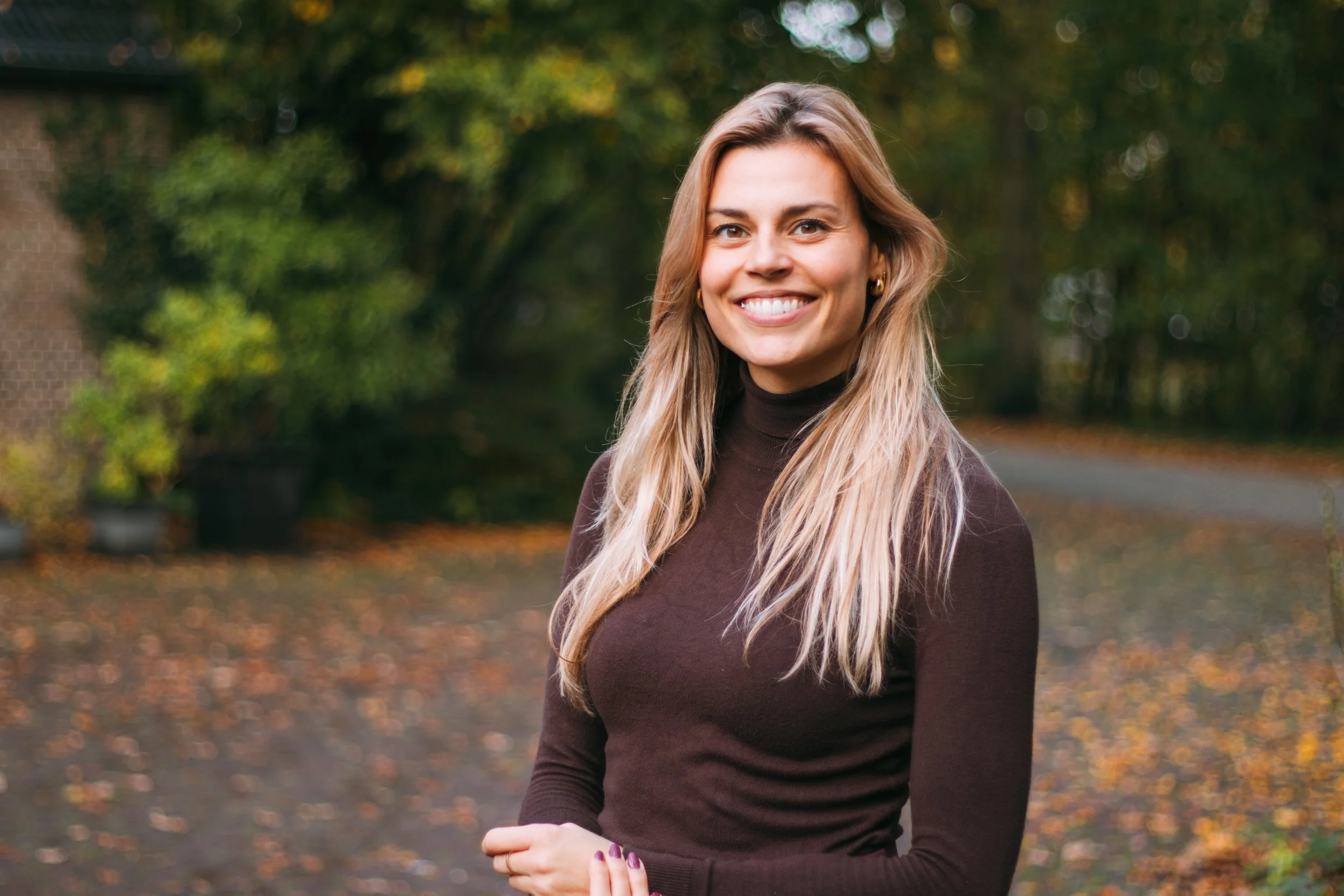 Vrouw met lange blonde haar glimlachend in een park met herfstbladeren en groene bomen op de achtergrond.