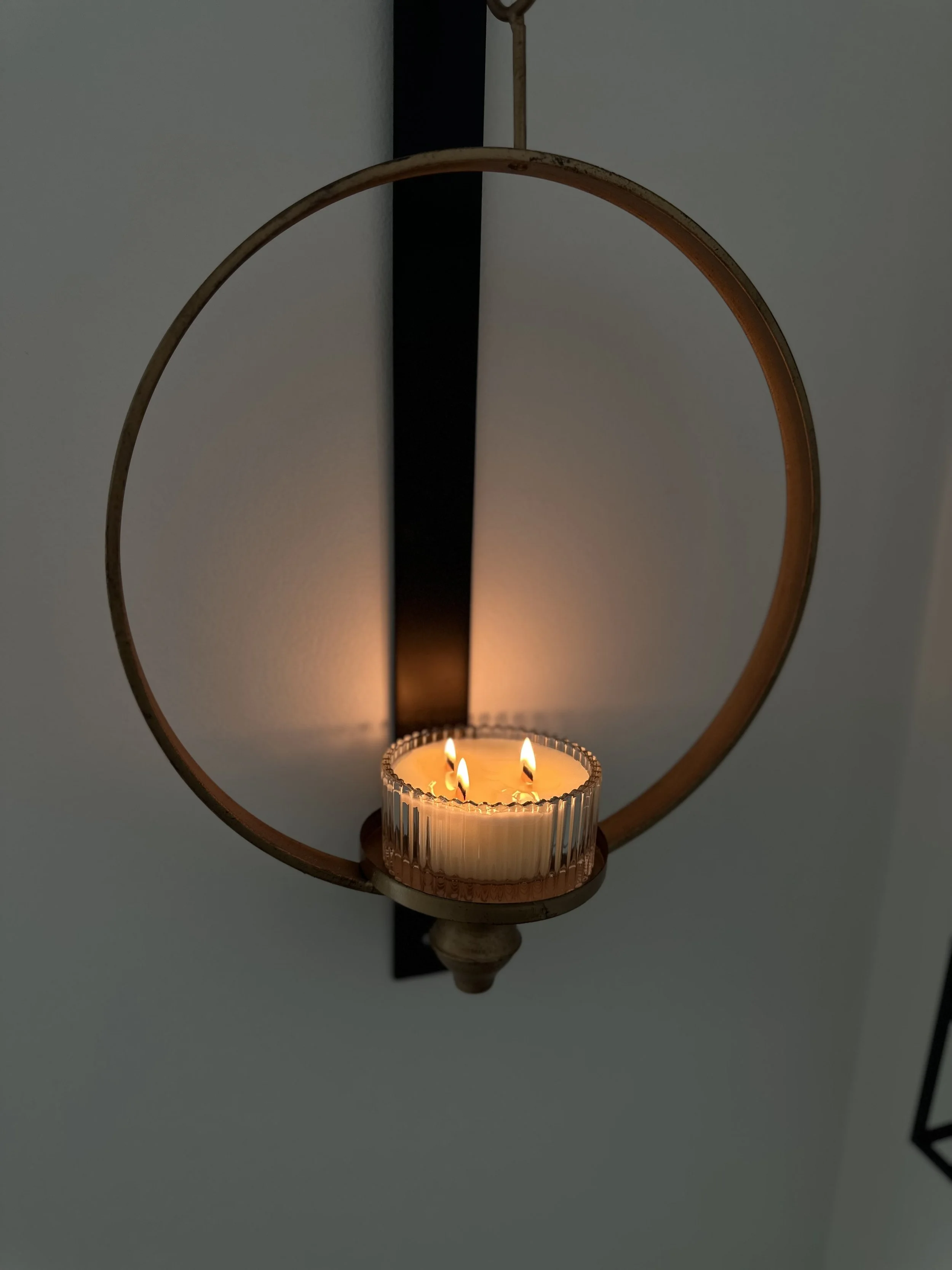 A lit candle with three flames inside a glass holder, mounted on a wall with a decorative circular metal frame.