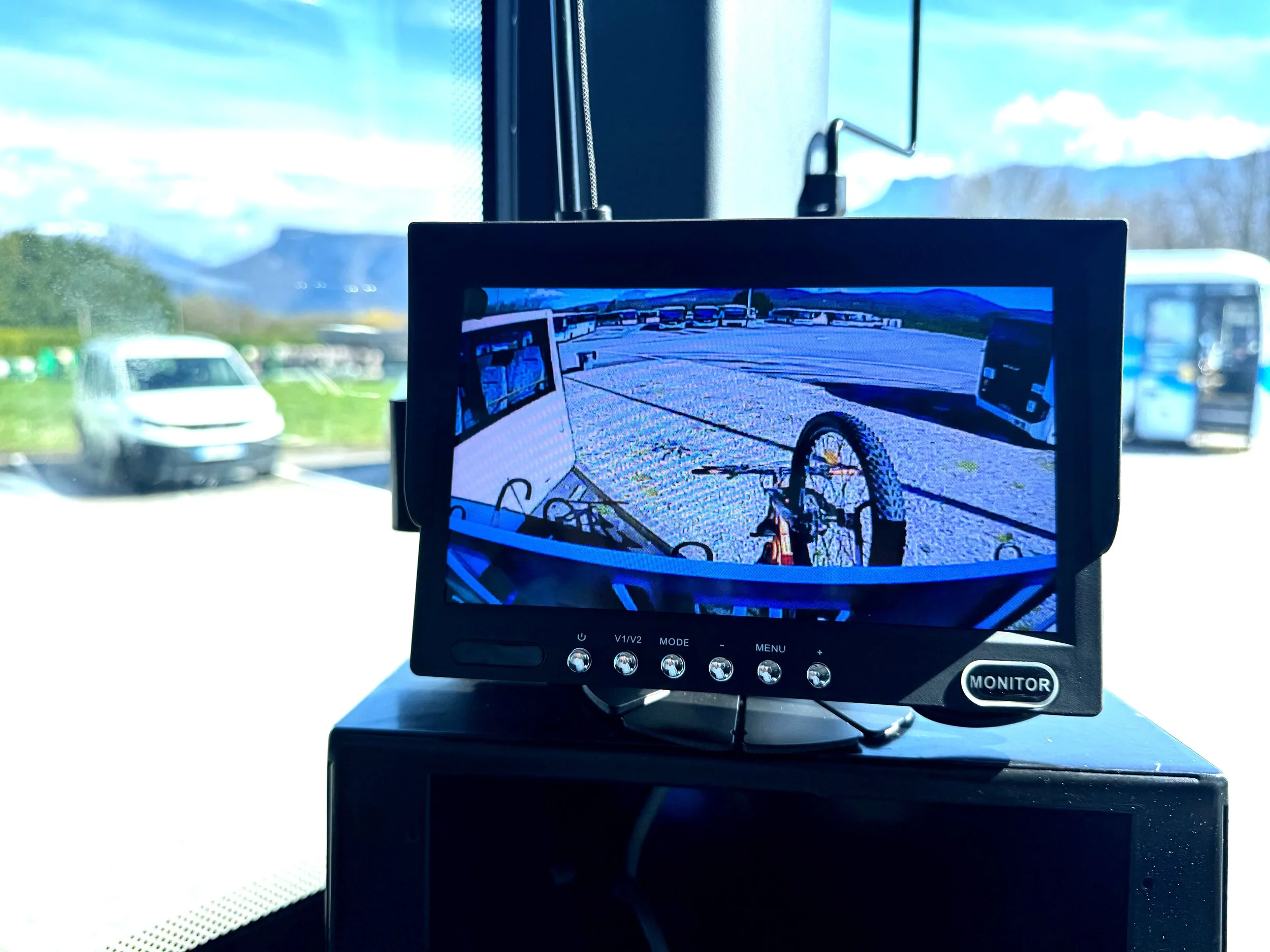 Écran de moniteur affichant la vue d'une caméra de recul, montrant un vélo à l'envers dans un parking avec d'autres véhicules et un arrière-plan de montagnes et de ciel bleu.