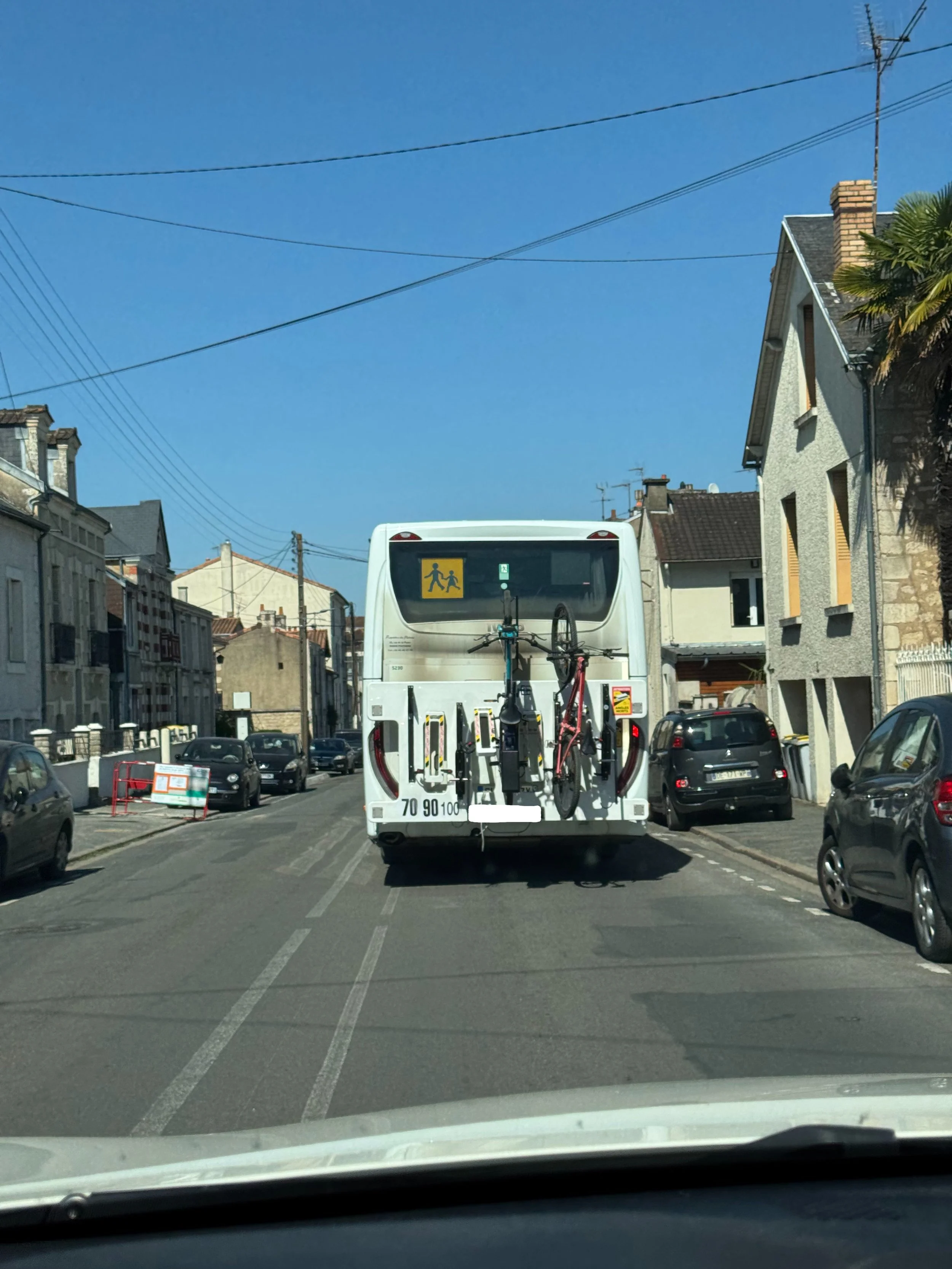 Un bus blanc avec deux vélos attachés à l'arrière, en circulation dans une rue urbaine, sous un ciel bleu.