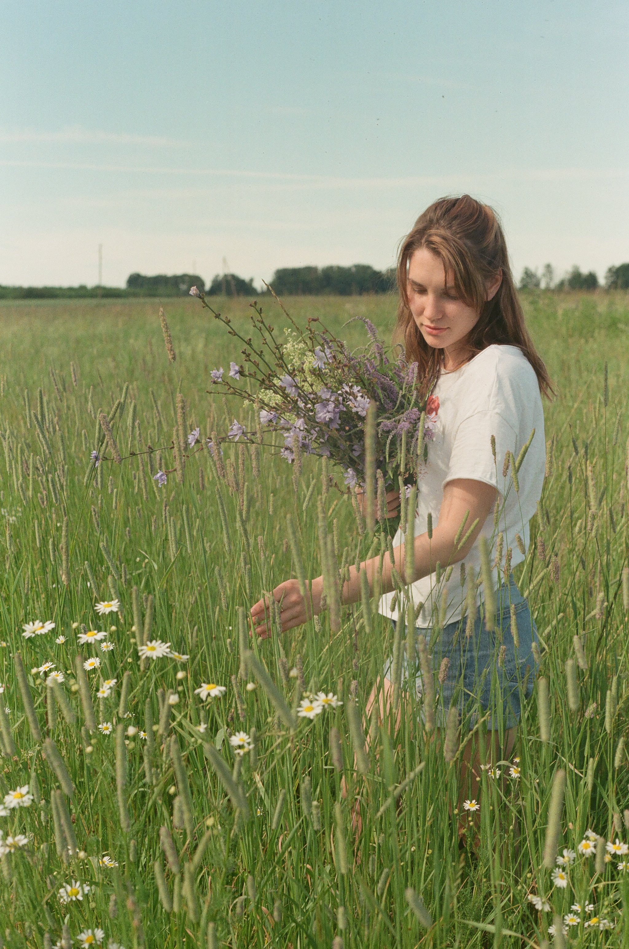 A young woman with light brown hair and fair skin is standing in a green field holding a bouquet of wildflowers, including purple and white blossoms. She is wearing a white t-shirt with a floral design and blue jeans, and is looking down at the flowers with a calm expression. The background features a wide open sky and distant trees.