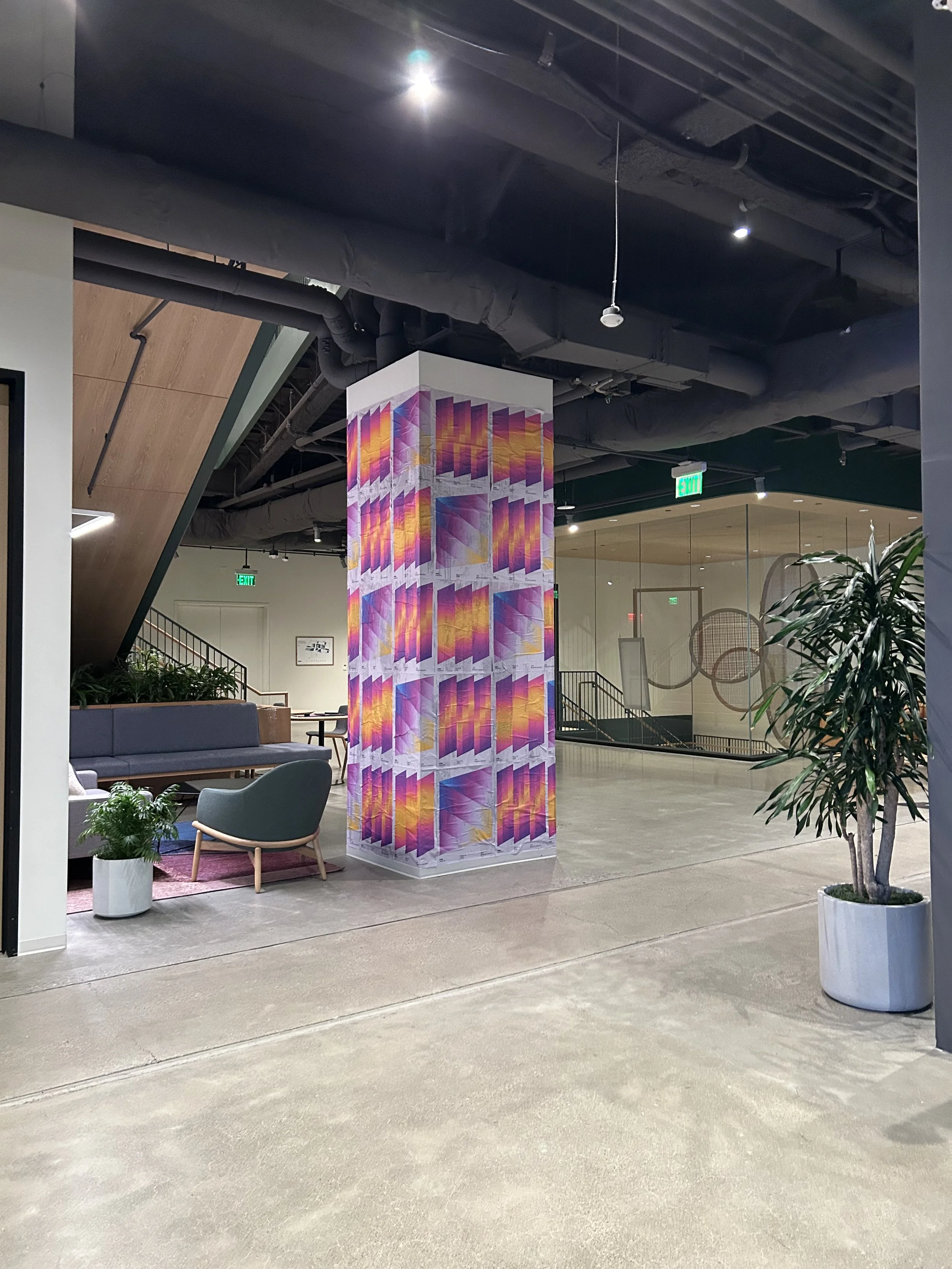 Interior of a modern building with a colorful abstract mural on a pillar, green plants, seating area, and exit signs.