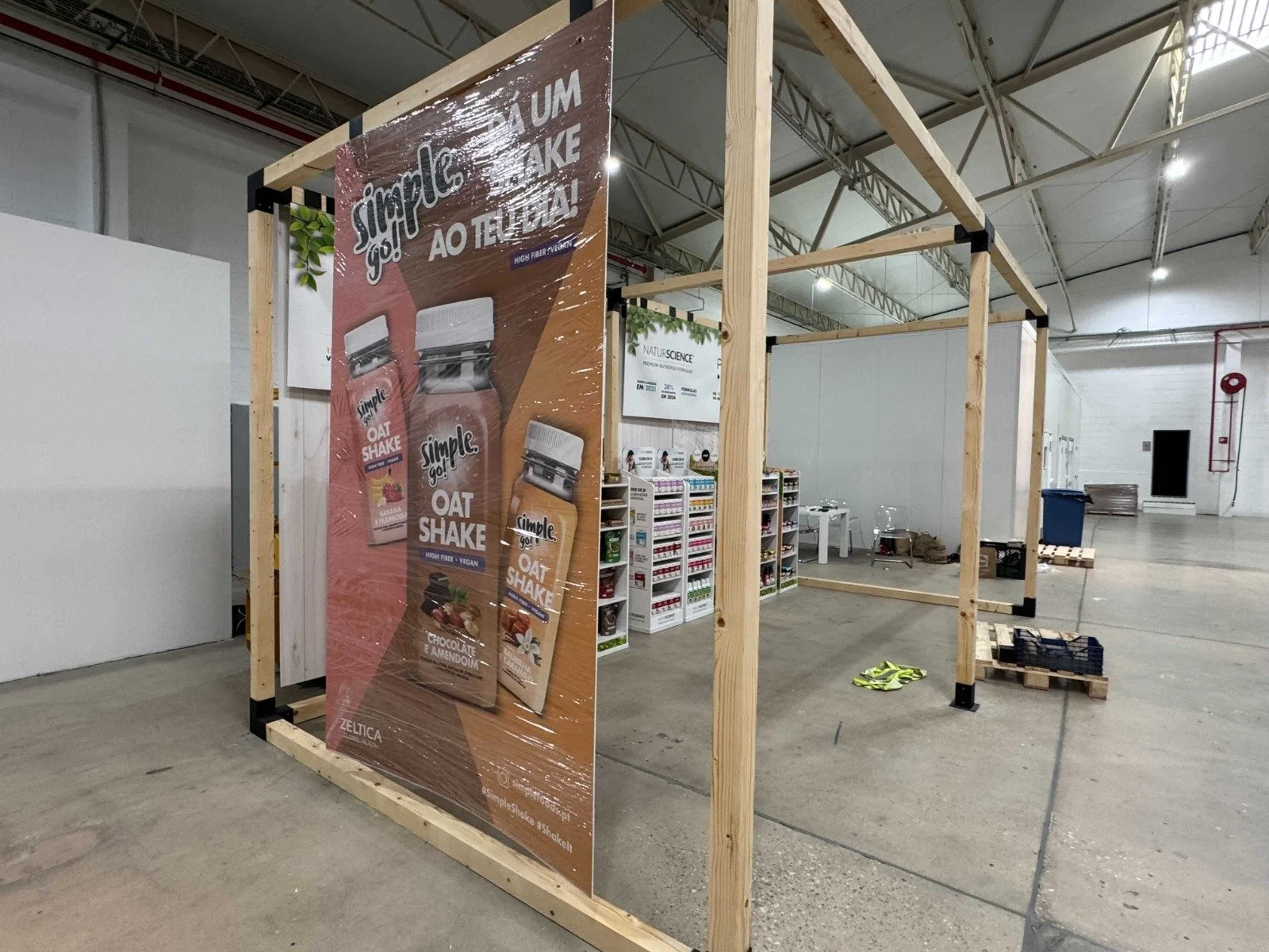 The image shows an indoor space under construction with a wooden framework. There is a large advertisement banner on the left side promoting a product called 'Simple Go! Oat Shake'. In the background, there are shelves filled with various packaged pr