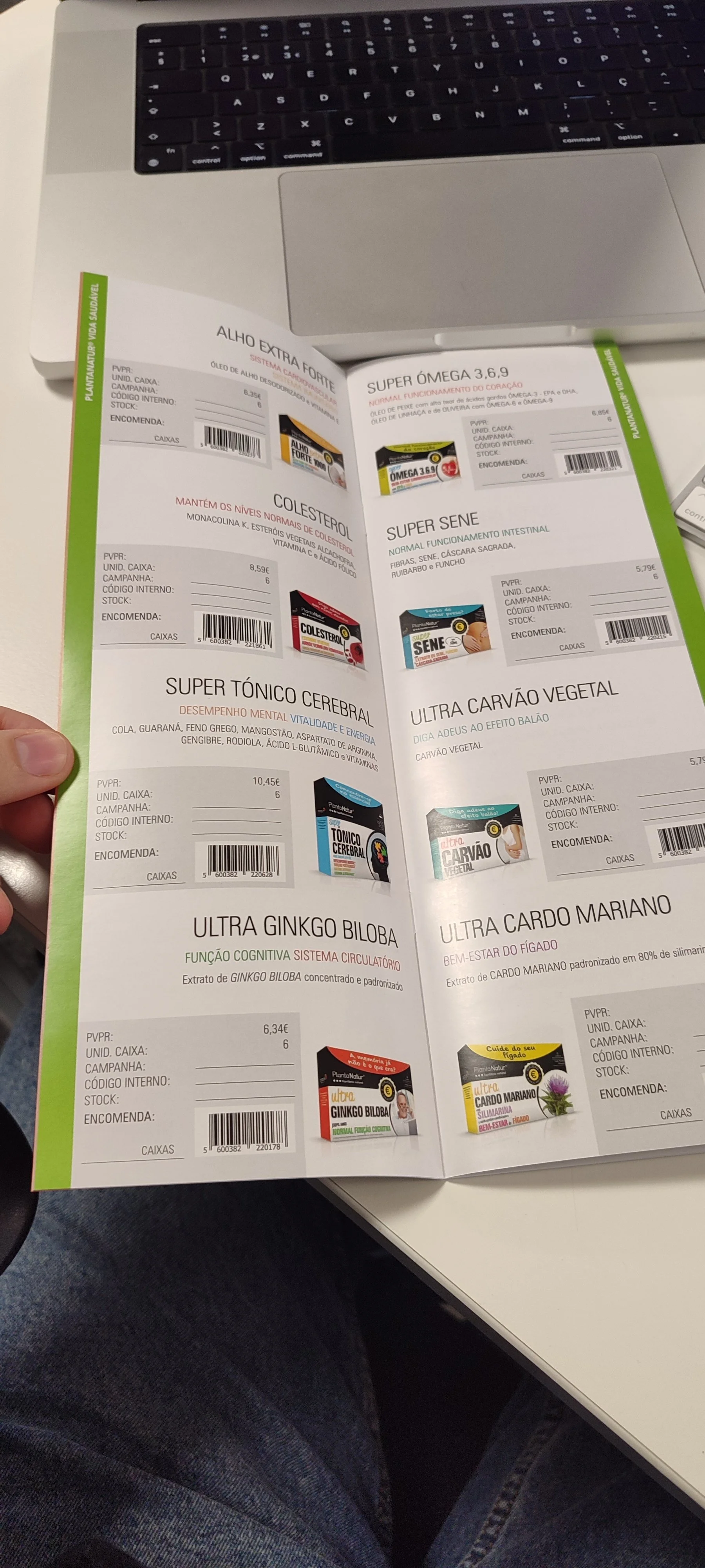 A person holding a brochure open on a white table, showing various health supplement products with descriptions and prices, in front of a laptop keyboard.