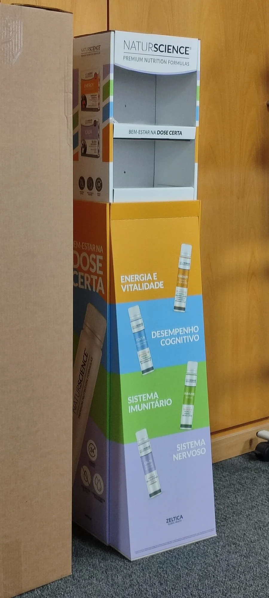 A cardboard display stand for NaturScience dietary supplements featuring colorful sections labeled with health benefits such as energy, cognitive performance, immune system, and nervous system, and a empty white shelf on top.