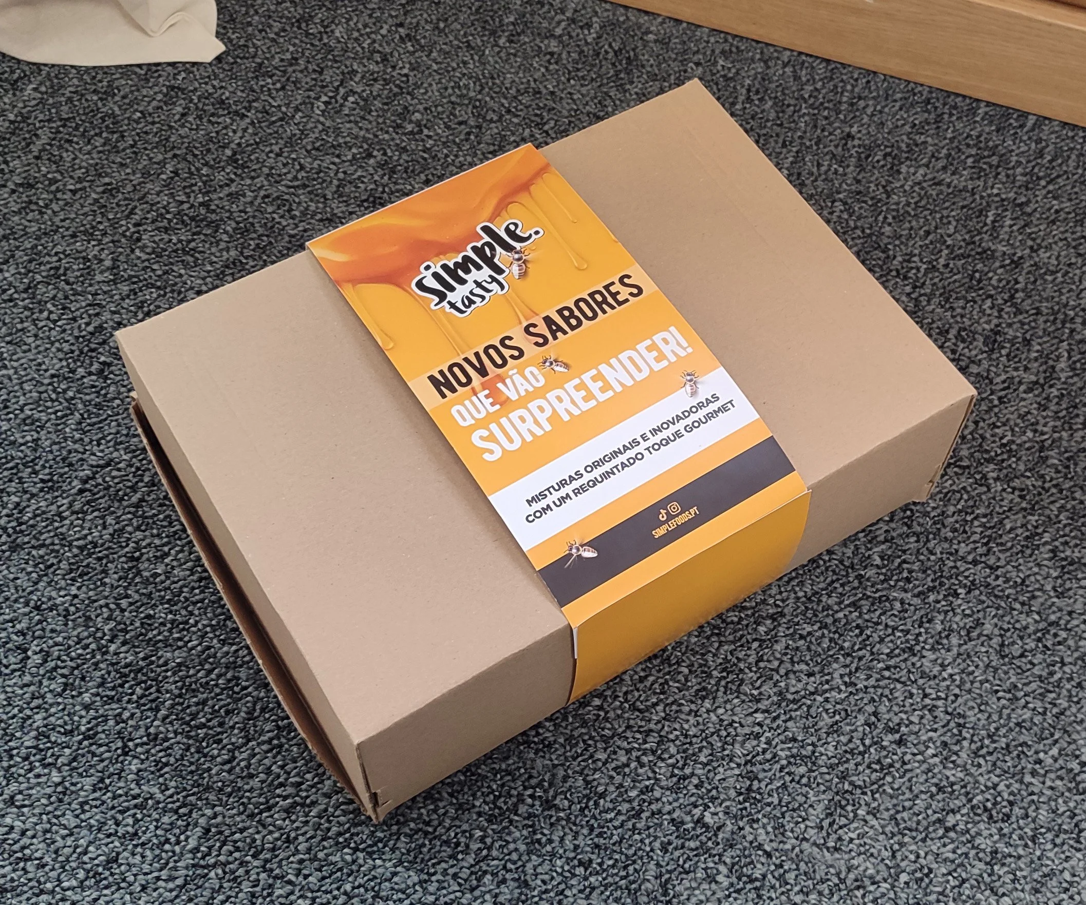 A closed cardboard box on a carpeted floor, with a colorful label featuring the brand 'Simple Taste' and text in Portuguese promoting new surprising flavors.
