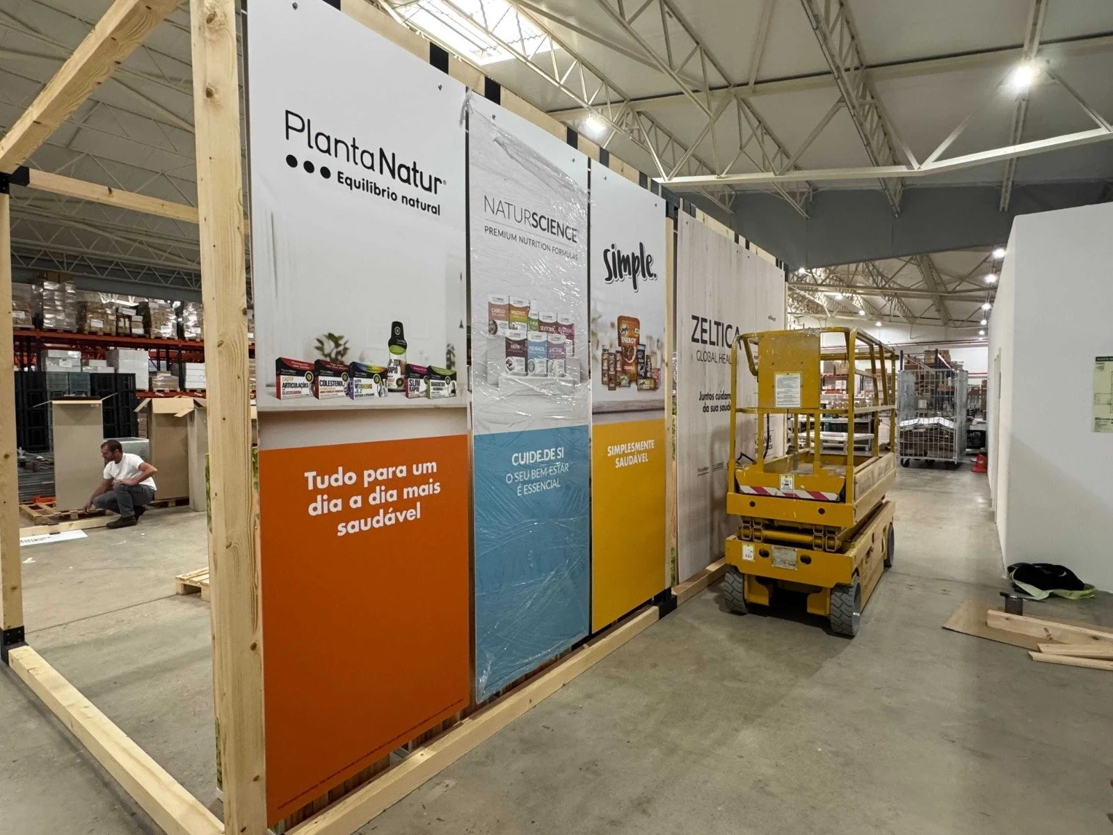 A construction site inside a warehouse shows a man working near a partially built wooden frame wall. There are large advertisement posters in Spanish on the wall, and a yellow scissor lift is parked next to it. The warehouse has a high ceiling with m