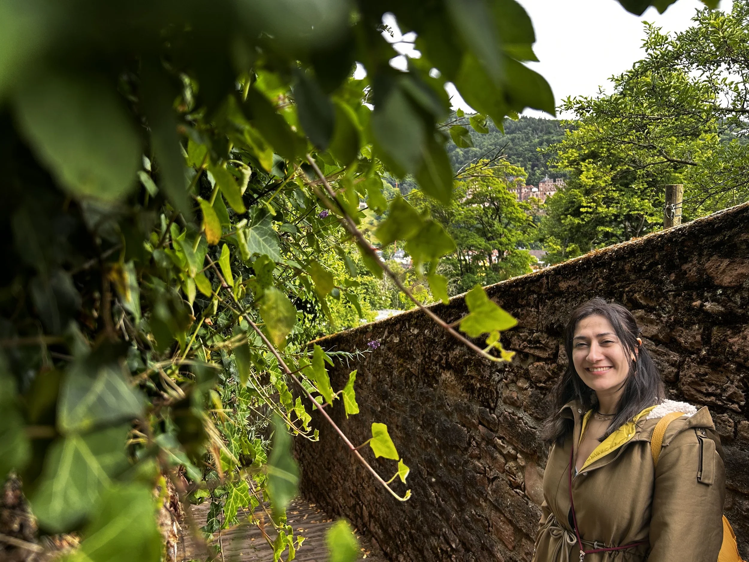 A woman smiling outdoors, standing beside a moss-covered stone wall with dense green ivy in the foreground. In the background, a hillside with trees and some buildings is visible.