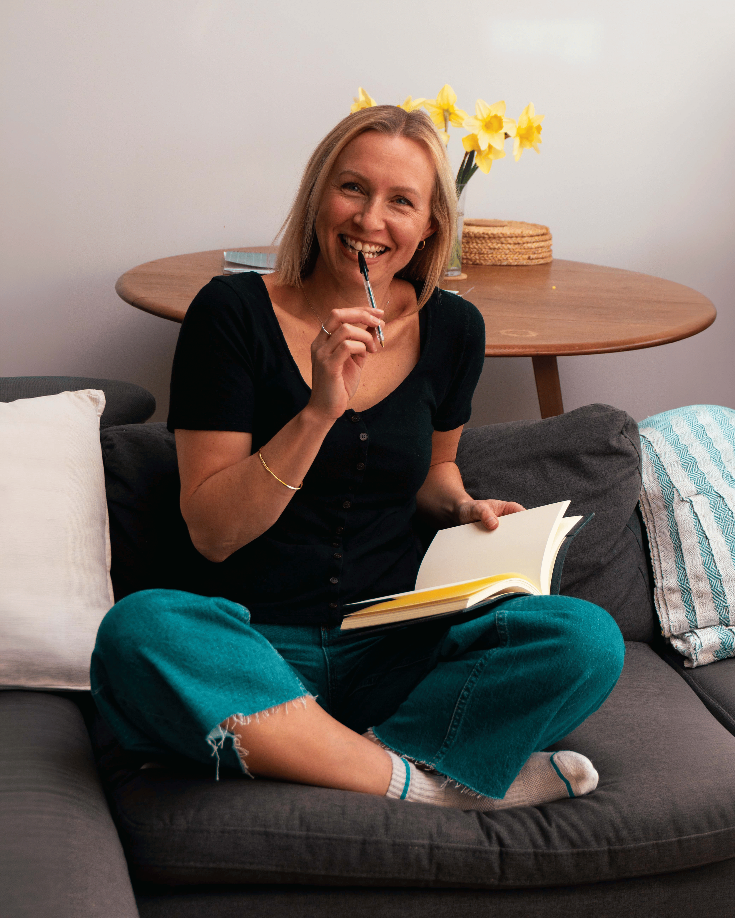 Rowan smiling and sitting on a couch, holding a notebook and a pen, with a yellow daffodil bouquet on a wooden table behind her and wearing casual clothes.