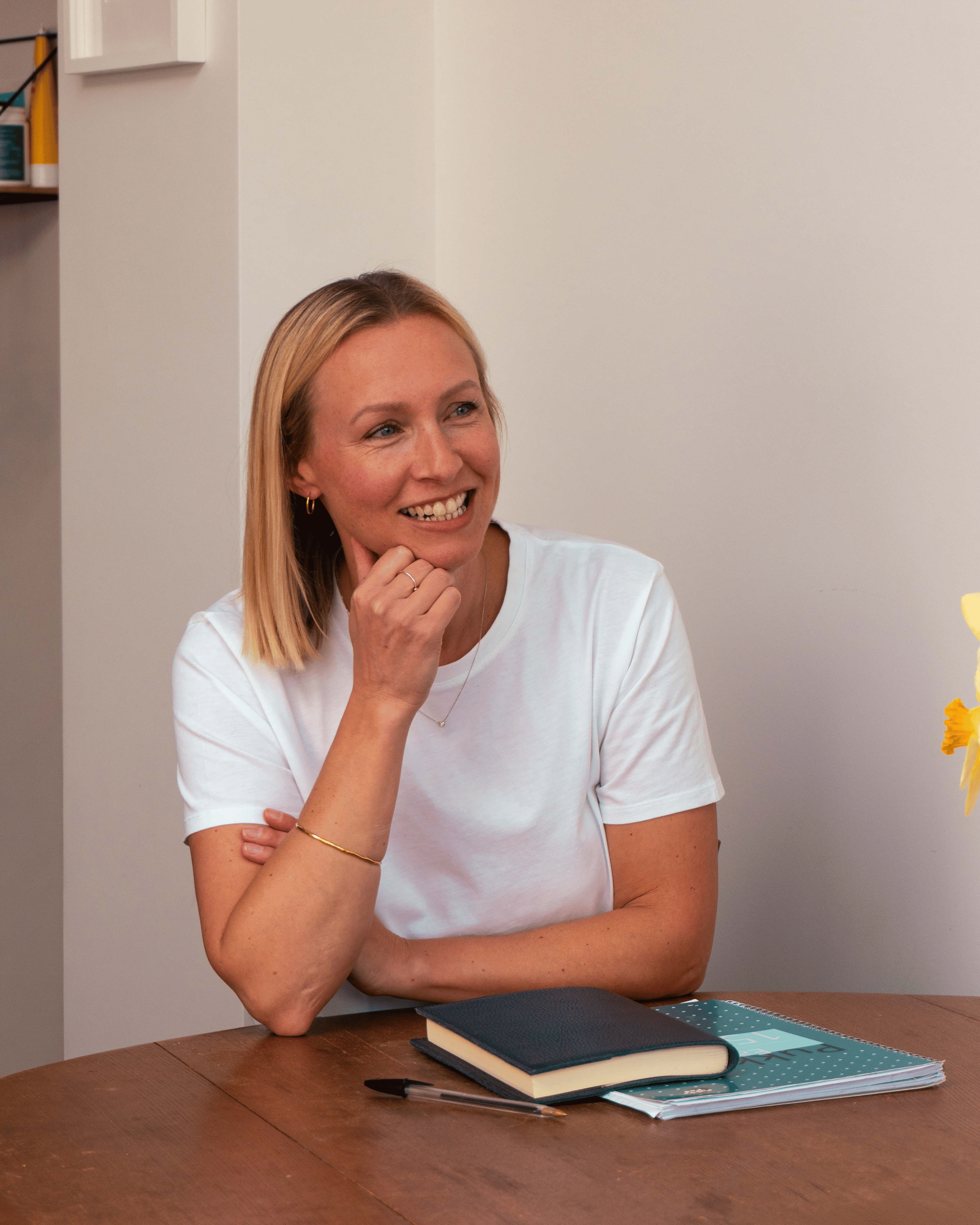 Rowan sitting at a wooden table with a smile, wearing a white t-shirt, with notebooks, a pen, and a pencil case on the table.