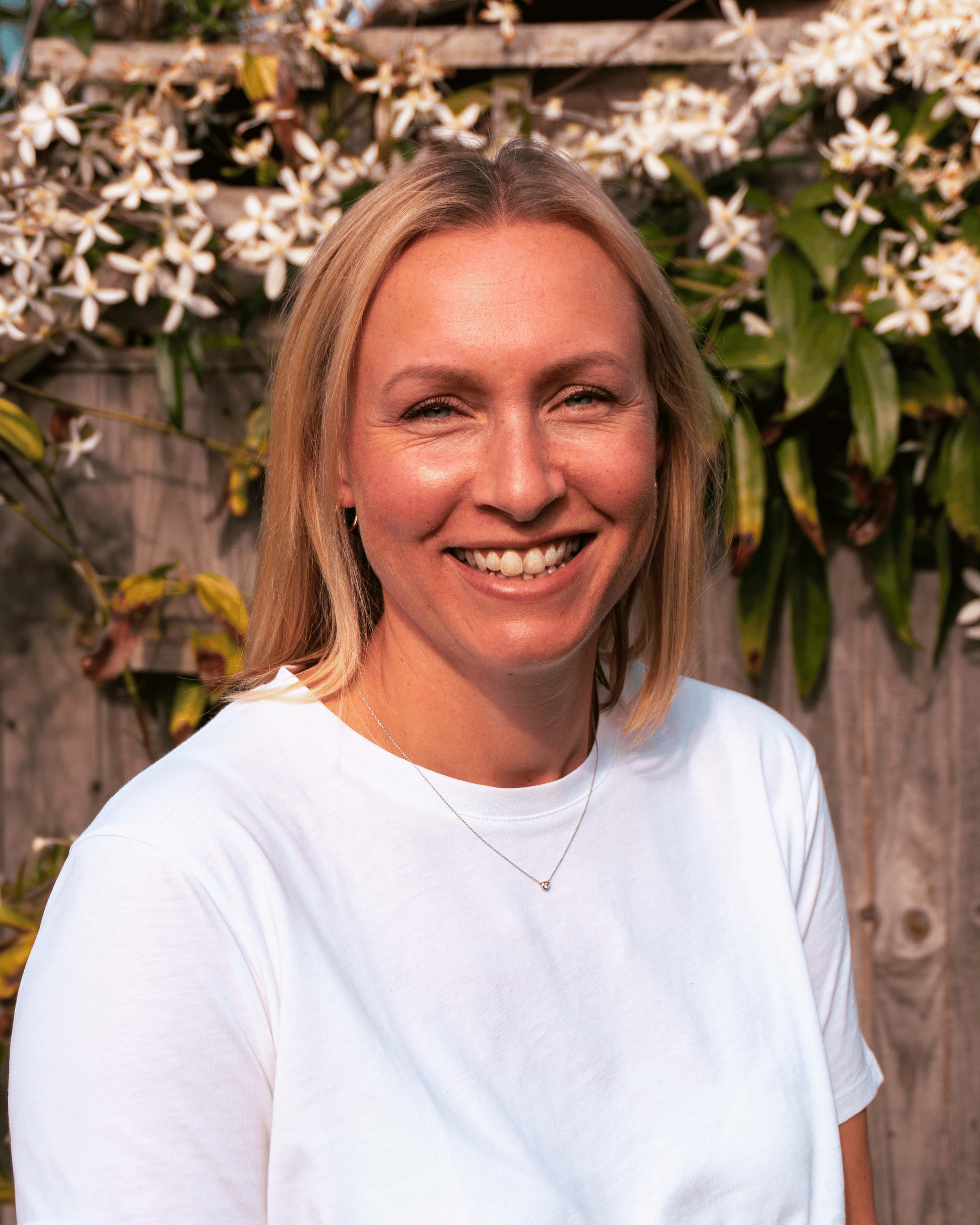 Rowan smiling outdoors in front of a wooden fence with blooming white flowers and green leaves.