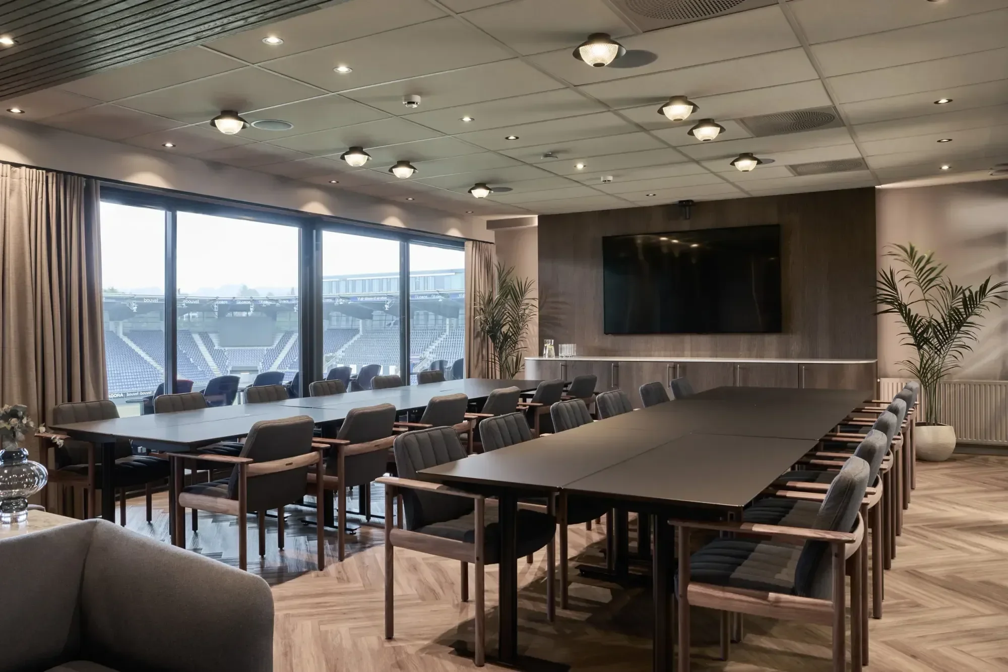 Interiør av et moderne konferanserom med store vinduer, langbord, stoler, en flatskjerm, samt planter.