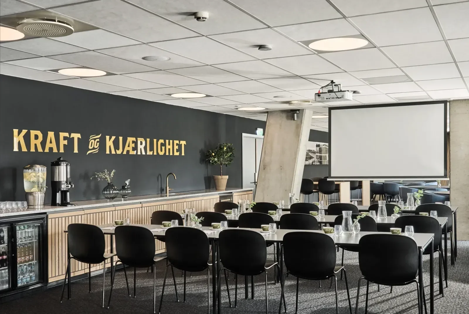 Et konferanserom med svarte stoler rundt hvite bord, dekorert med vaser og glass, samt et stort lerret og en vegg med teksten "KRAFT og KJÆRLIGHET"