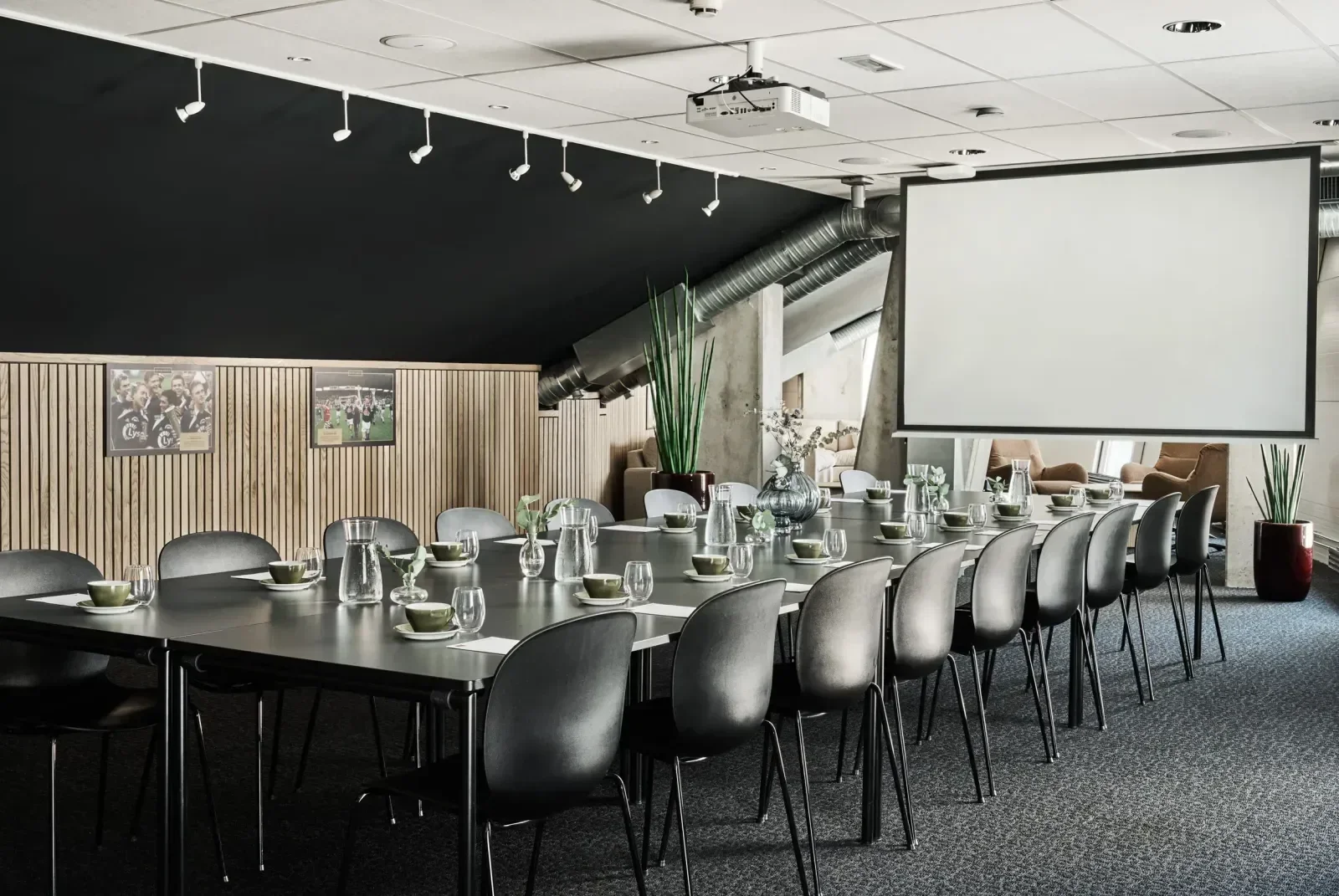 Konferanserom med lang svart bord, svarte stoler, store vaser med grønne planter, bilder på veggen, projeksjonskjerm og naturlig lys.