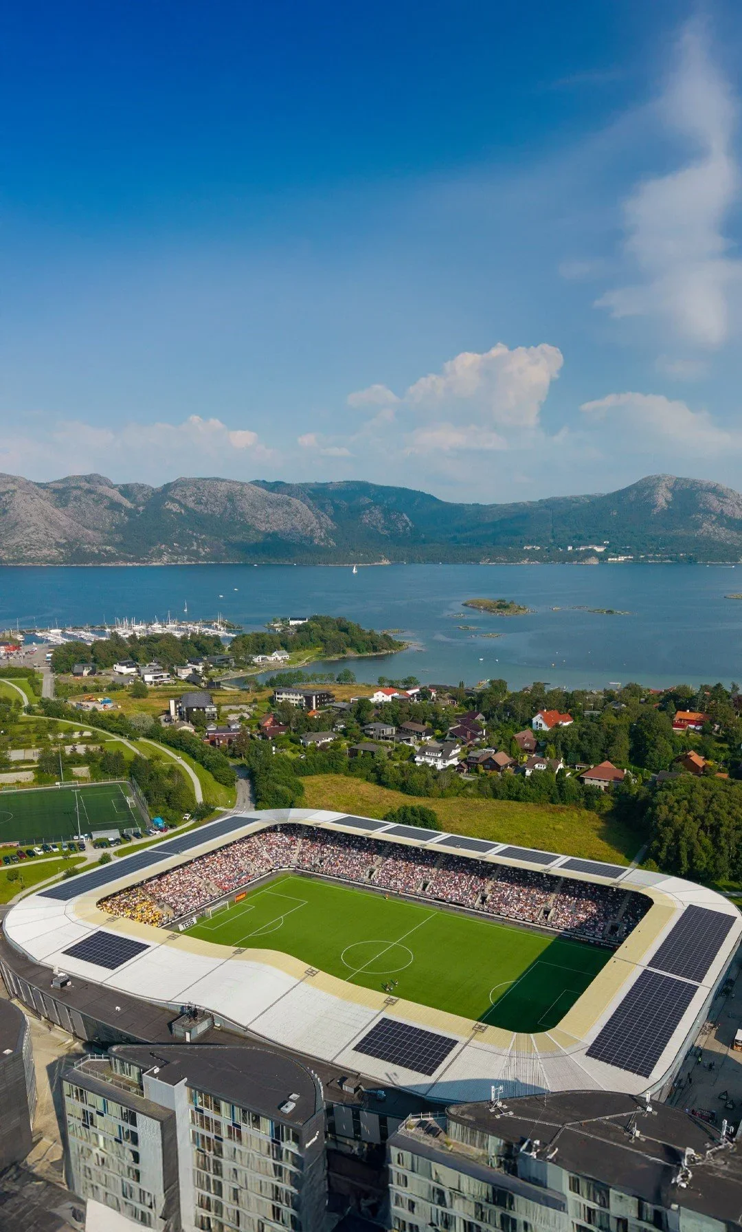 Landskap med en fotballstadion i forgrunnen, sjø og øyer i midten, fjell i bakgrunnen, blå himmel med noen skyer.