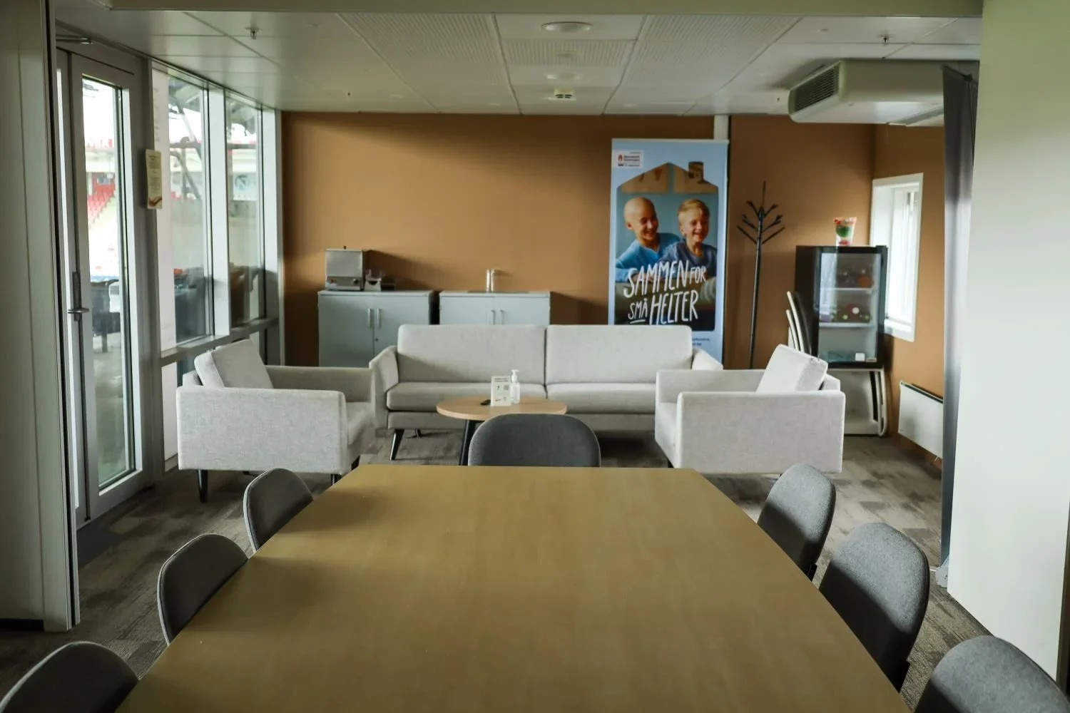 Et lite møte- eller venterom med et tregulv, et bord med sju stoler rundt, en hvit sofa, en koffert, en bilderamme på veggen, en klesstativ, et lite skap, et vannfontene og noen vinduer.