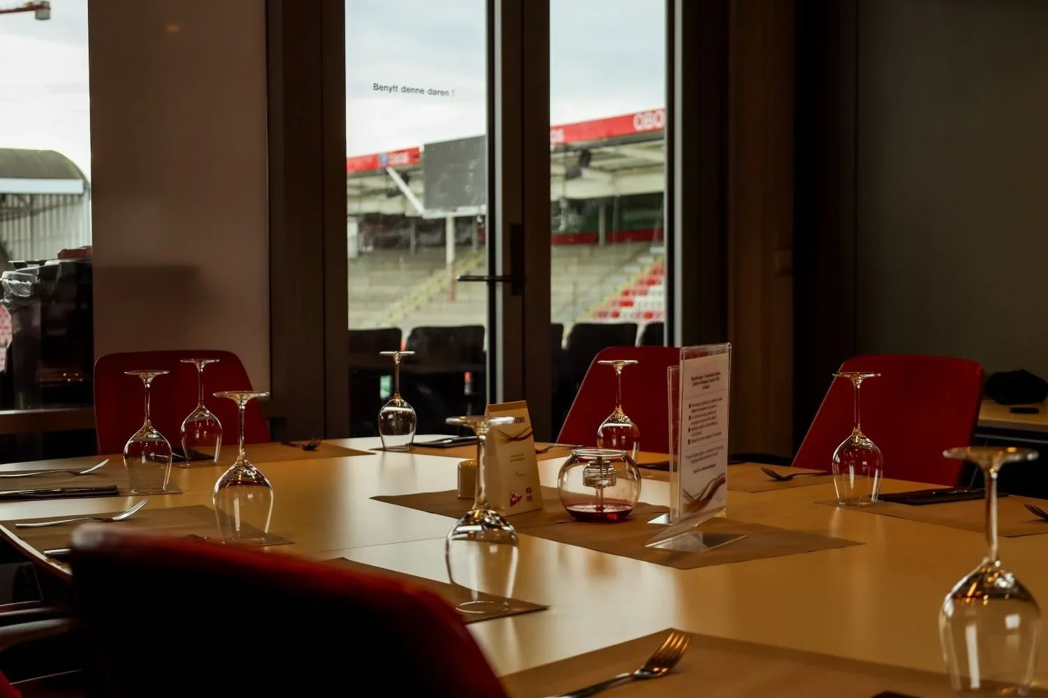 Innendørs restaurantbord med tomme glass, bestikk og menyer, med utsikt til et utendørs stadium gjennom store glassdører.