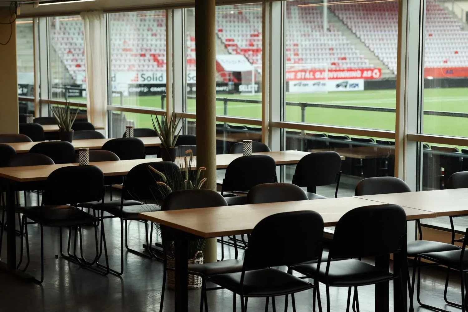 Innendørs utsikt over tomt ganske tomt rom med svarte stoler og bord, store vinduer med utsikt over en fotballbane.