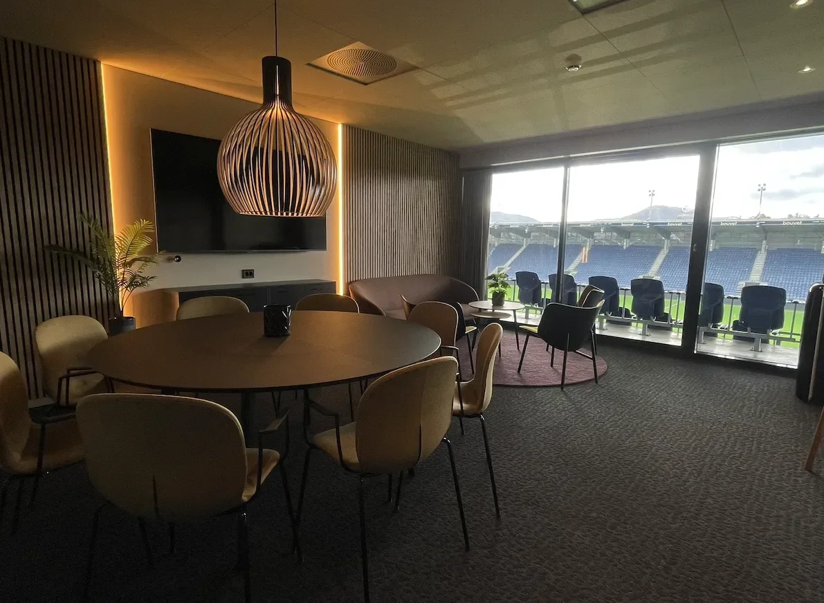 Innendørs rom med rund bord, stoler, sofa, store vinduer som viser en fotballstadion, og moderne belysning.