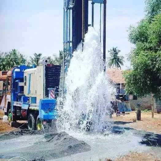 Camión realizando una perforación profunda en la tierra, expulsando agua y polvo en un entorno rural con árboles y casas.