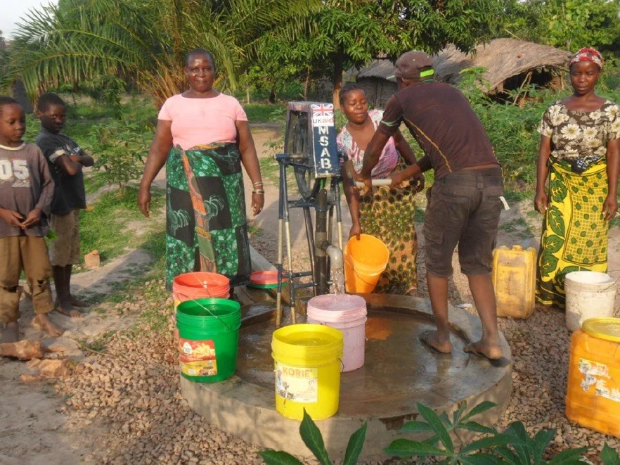 Personas en una estación de agua comunitaria sirviendo líquido en recipientes coloridos en un entorno rural con vegetación y casas de madera.