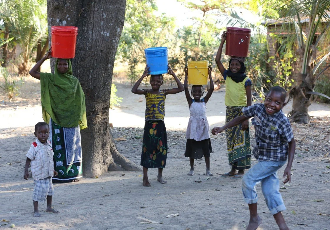 Grupo de niños y mujeres africanos sonriendo y levantando baldes de colores en un entorno al aire libre.
