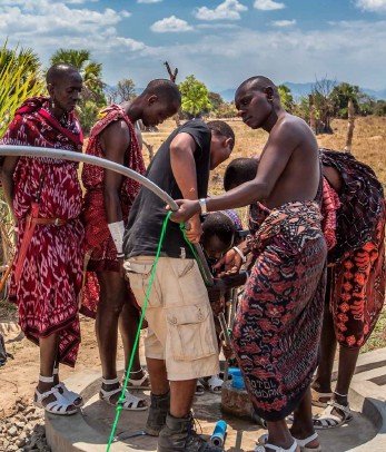 Grupo de personas, incluidos niños, bebiendo agua de un grifo al aire libre en un entorno rural.