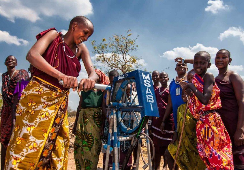 Mujer africana colocando una placa en una bicicleta en un campo con árboles y un cielo nublado, rodeada de niños y otras mujeres.