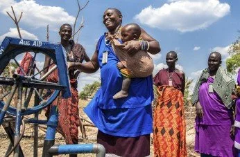 Un grupo de personas sonrientes, incluyendo una mujer con un niño, en un entorno al aire libre con un marquito de agua en primer plano y cielo azul con nubes.