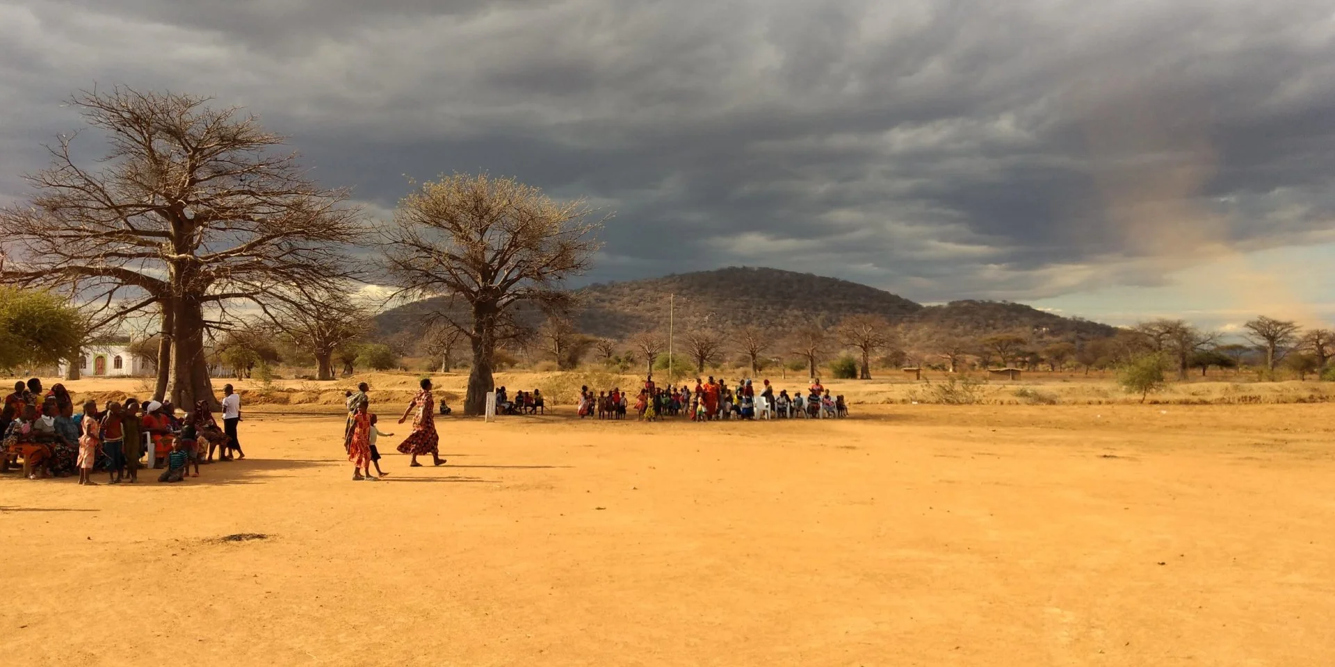 Grupo de personas africanas reunidas en un campo seco con árboles y montañas al fondo, cielo nublado. África.
