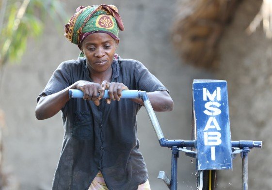 Mujer usando una camiseta gris y un turbante, operando una máquina de agua en un entorno exterior.
