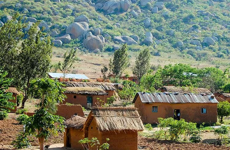 Vista de un pequeño pueblo africano con casas de barro y techos de paja, rodeado de árboles y un paisaje montañoso con rocas grandes en el fondo. África.