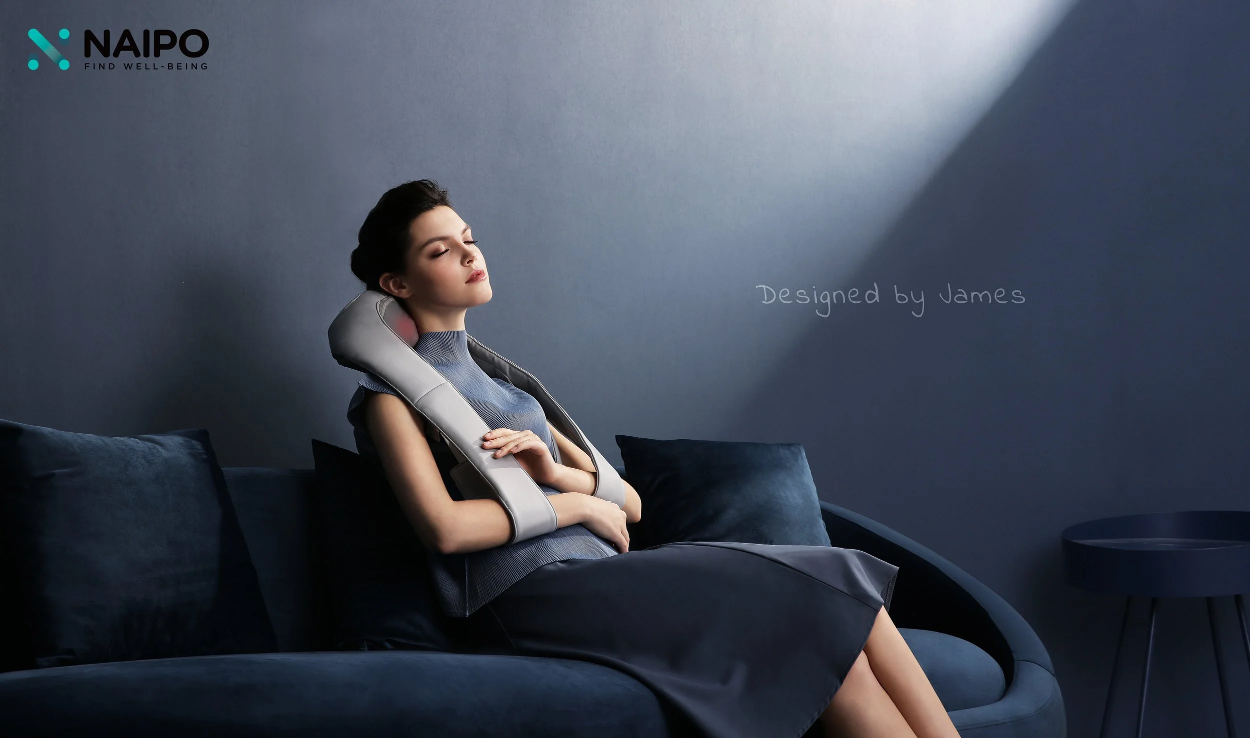A woman sitting on a dark blue couch with pillows, wearing a gray dress, with a massage neck pillow, eyes closed, relaxing in a dimly lit room with a gray wall and a small round table. Text on the wall says "Designed by James." Logo on top left corne